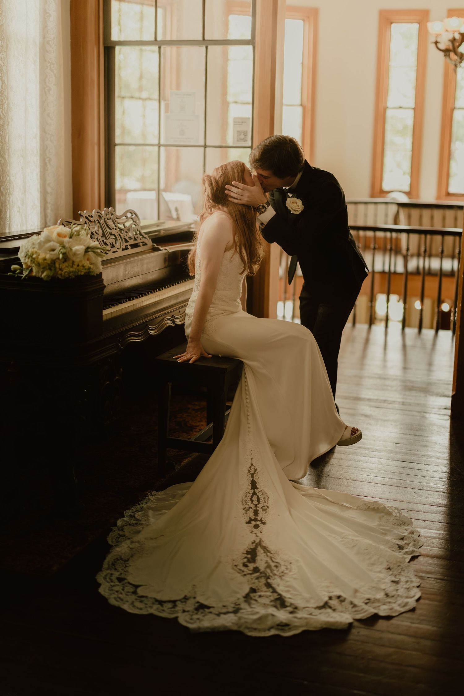 Bride in lace gown and groom in black suit share a romantic kiss by a vintage piano in a warmly lit venue.