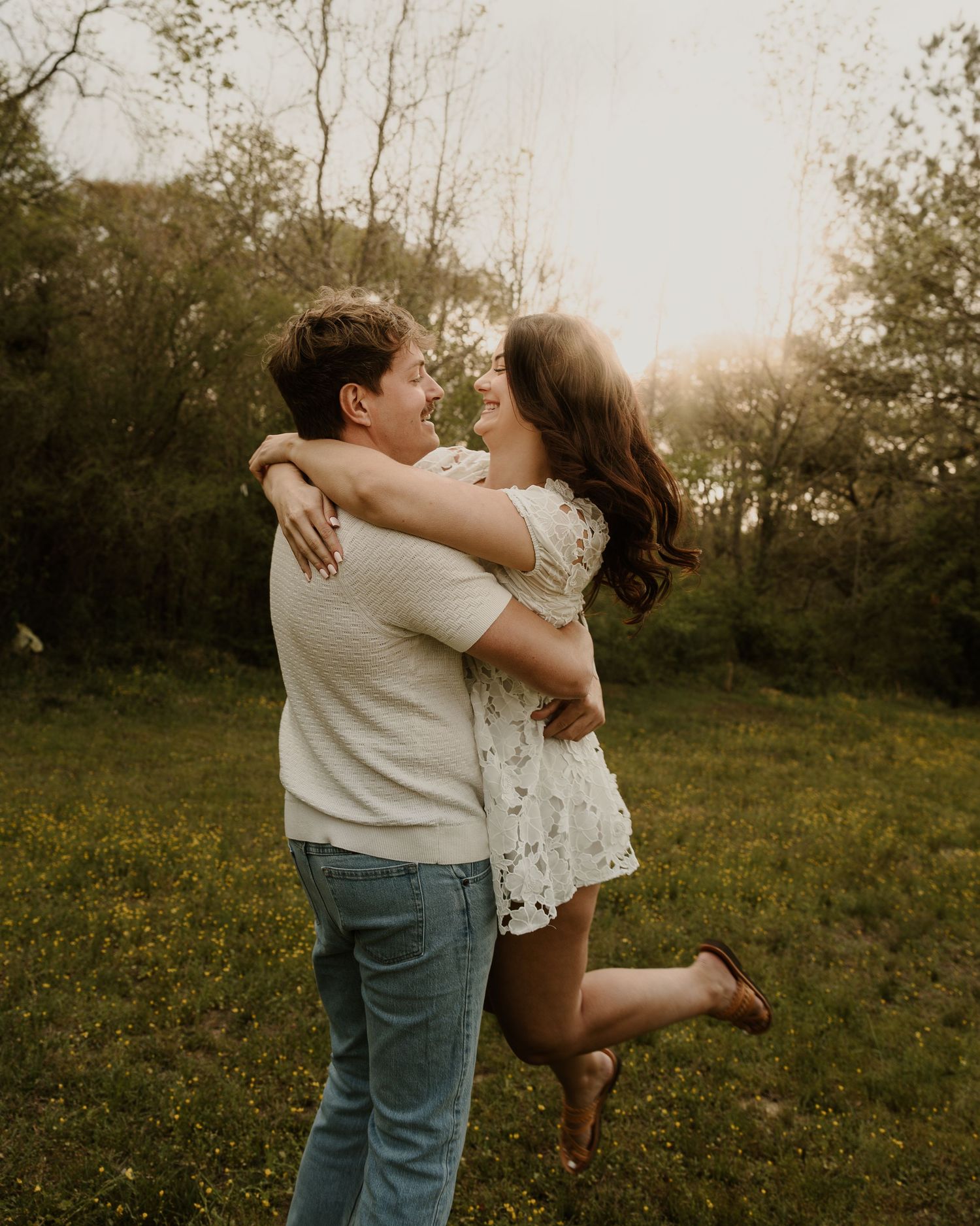 Couple embraces joyfully in sunlit meadow, man lifts woman in white lace dress, golden hour.