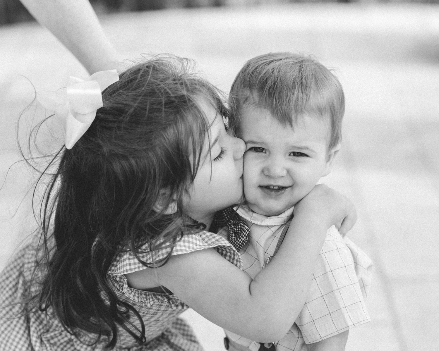 A young girl gives her smiling baby brother a sweet kiss on the cheek in a black and white photo.