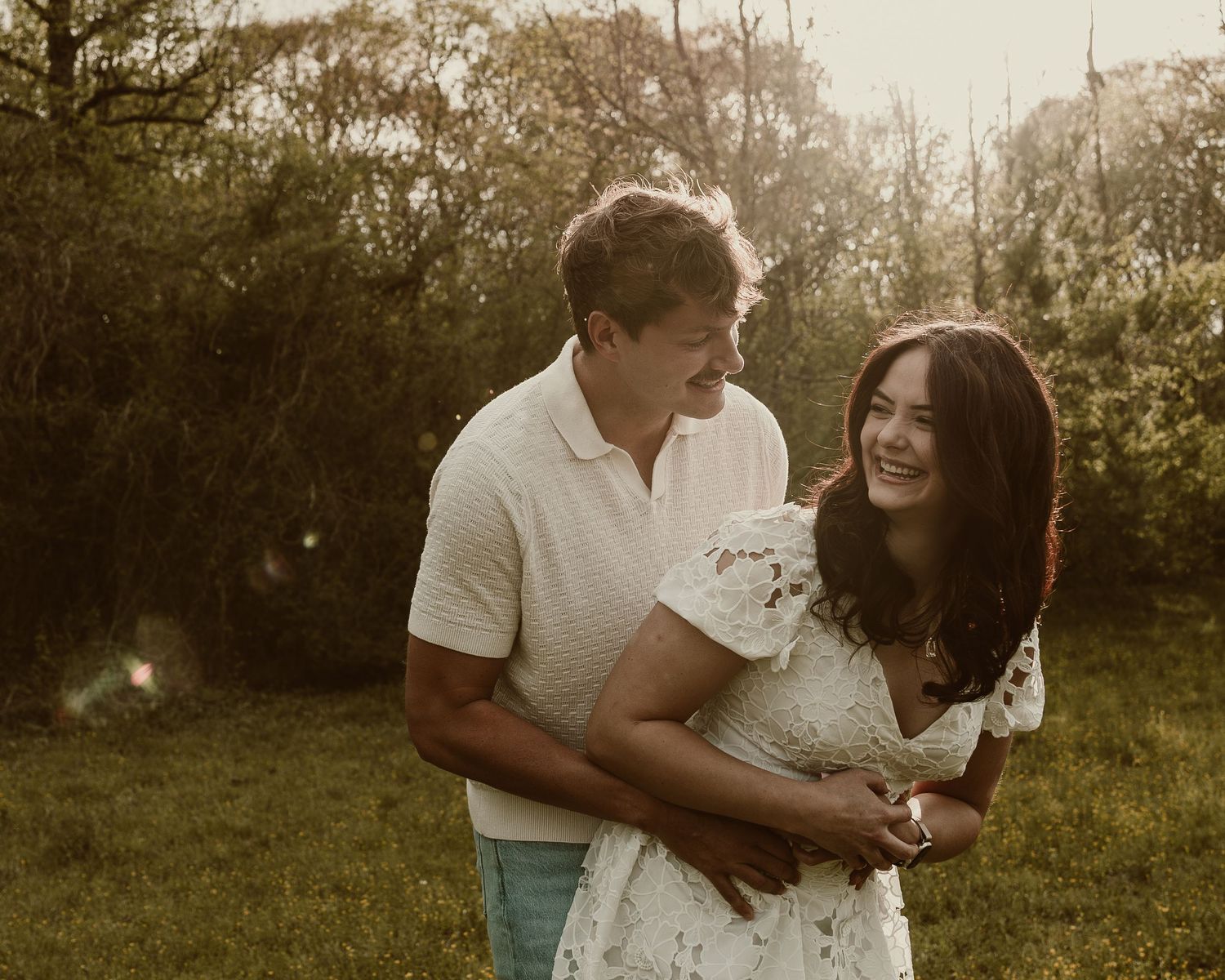 Happy couple embracing outdoors in warm golden light surrounded by lush green trees during engagement session.