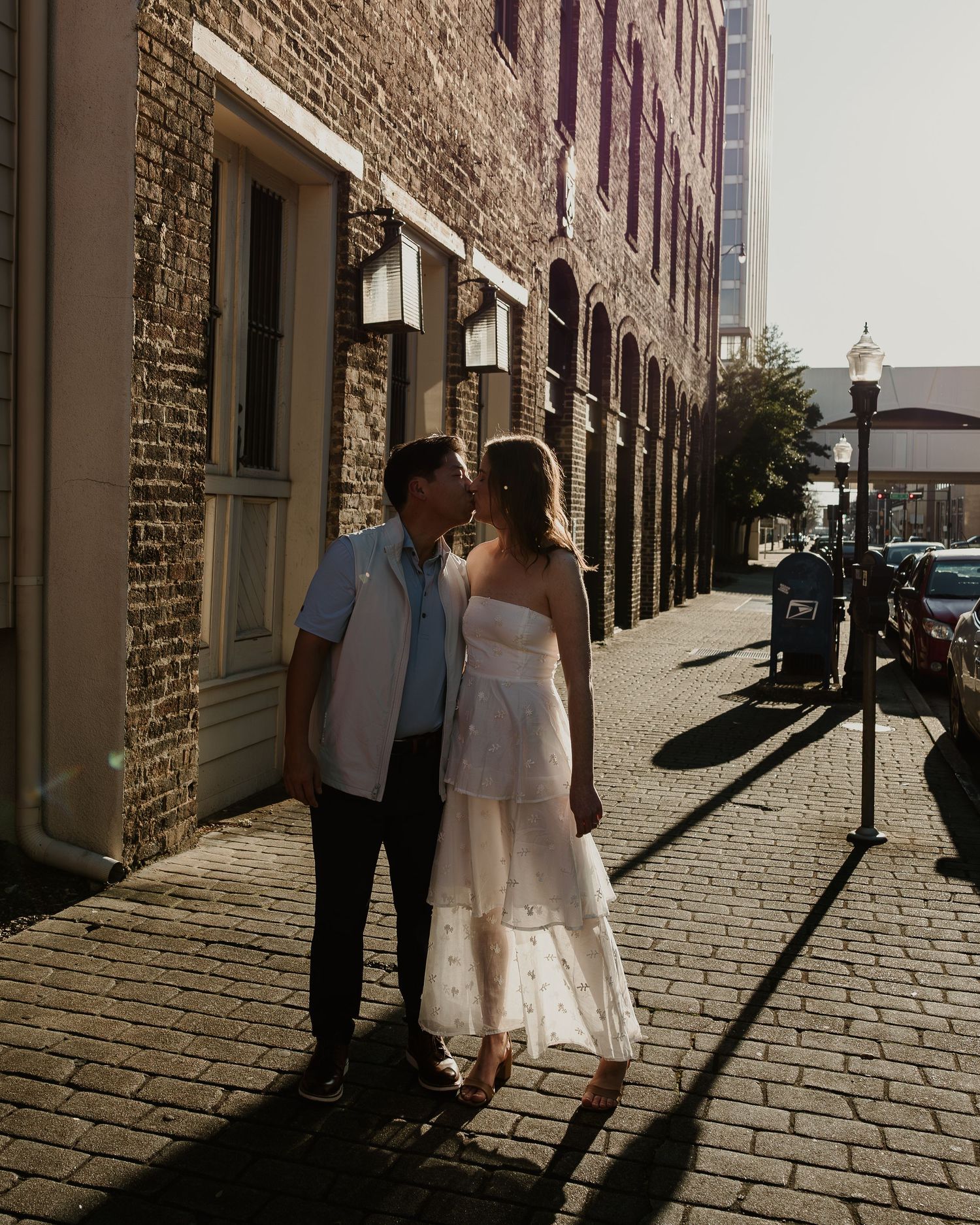 A couple in formal attire walks along a brick-lined alley casting long shadows in dramatic sunlight.