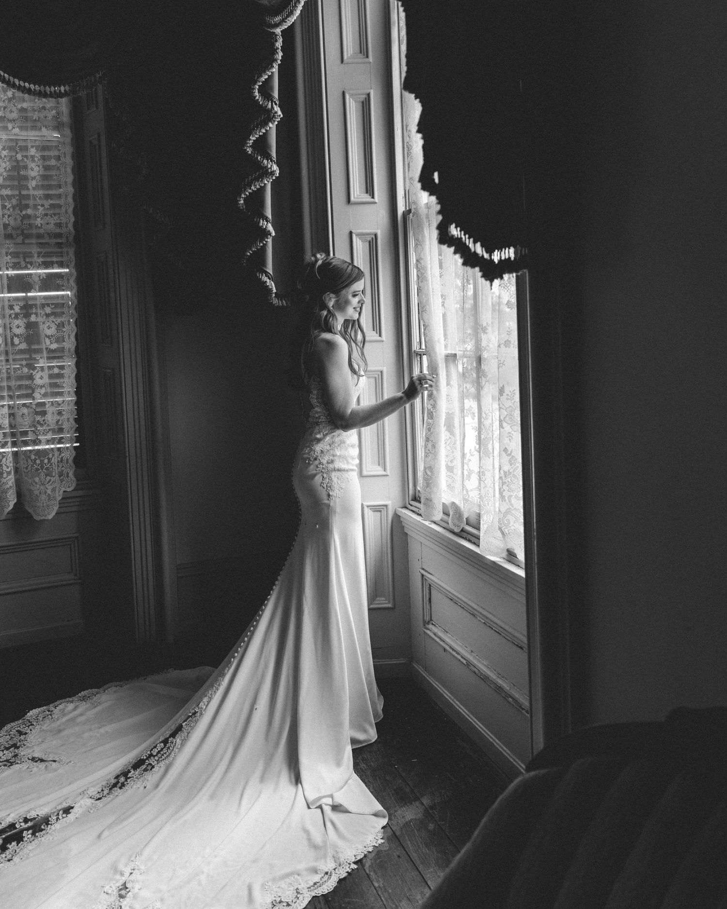 Black and white photo of a bride in a mermaid gown standing by a tall window, her lace train flowing behind her.