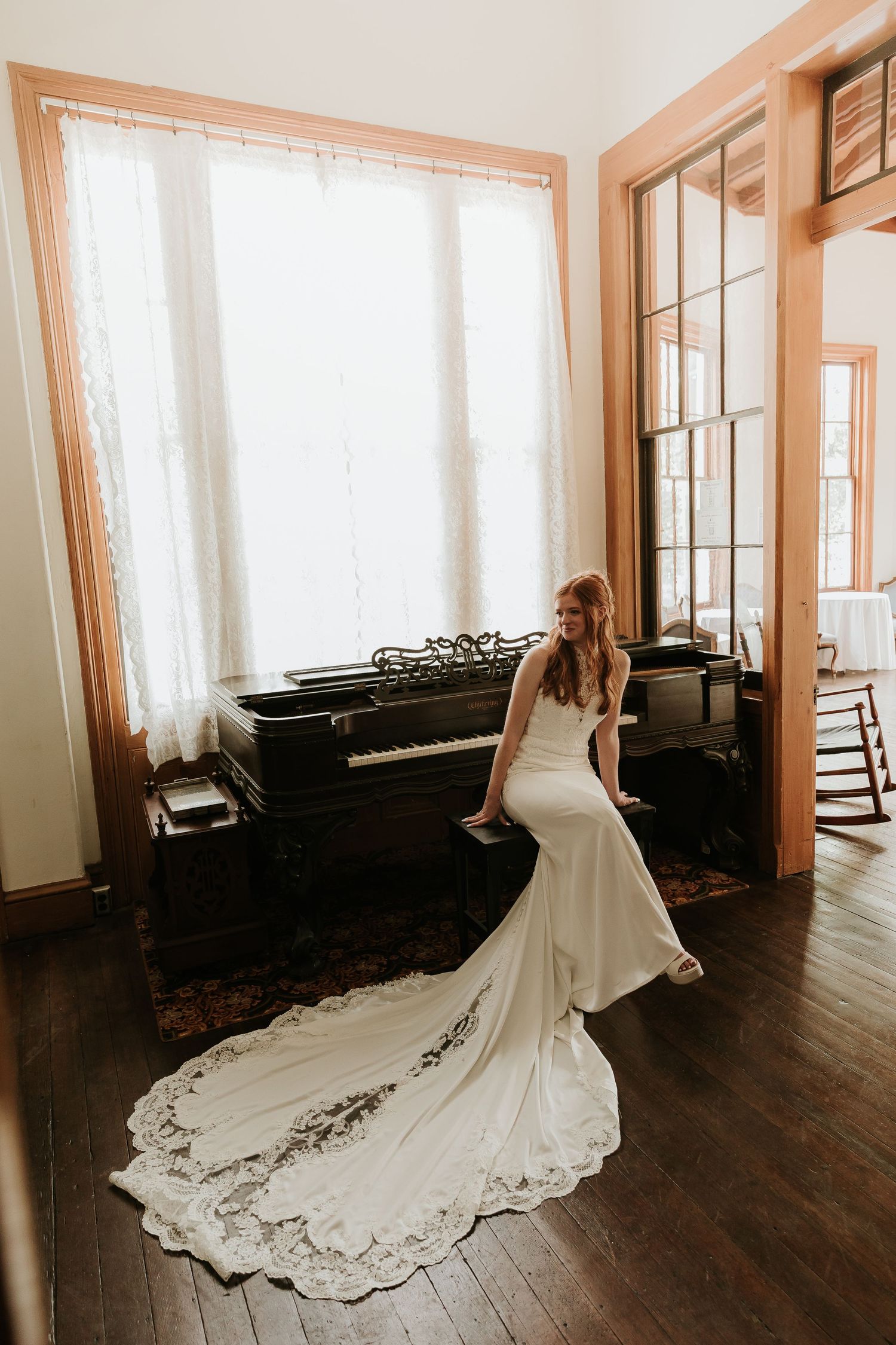 Bride in sleek mermaid wedding dress with lace train posing near grand piano in sunlit room.