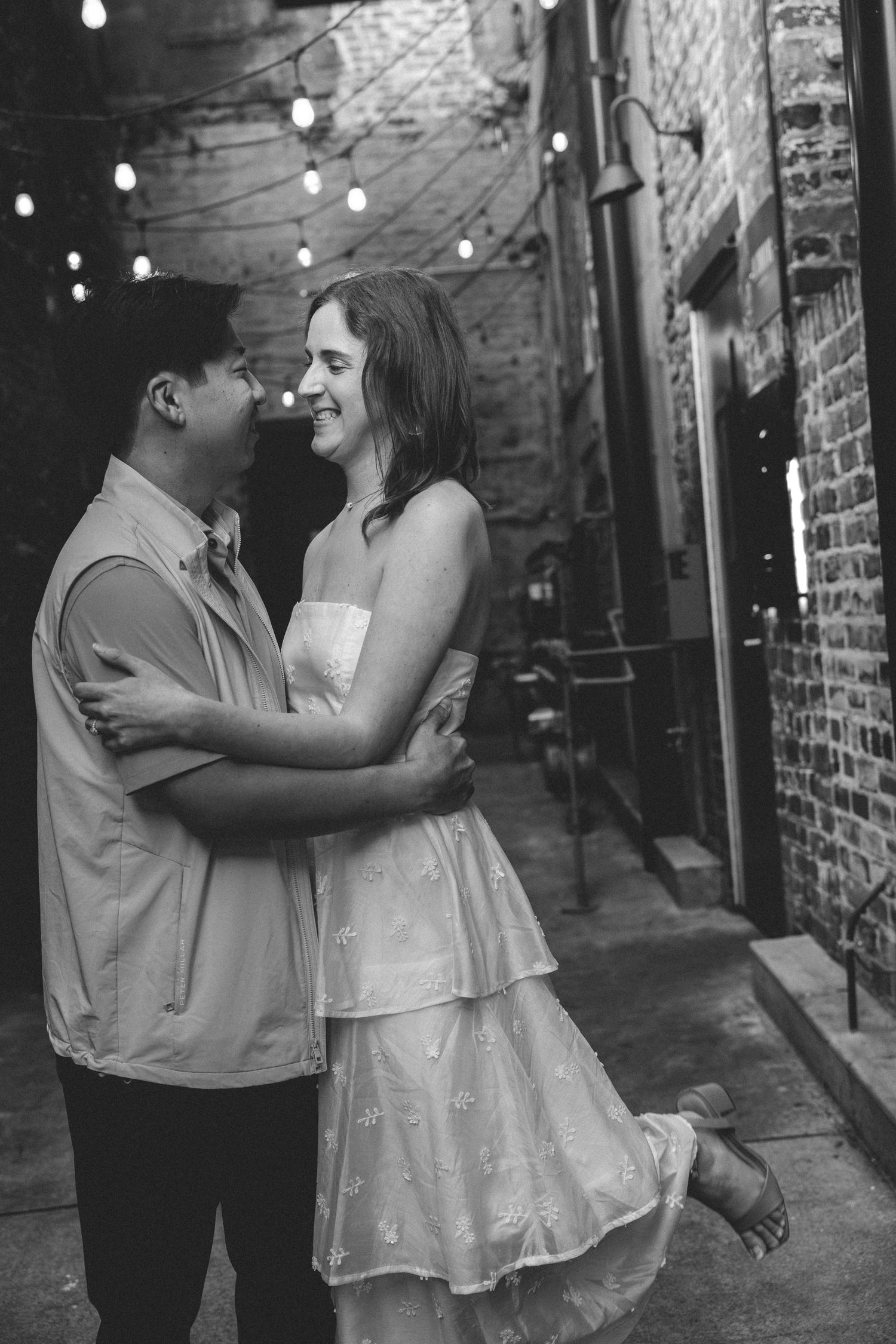 A romantic couple embraces in a dimly lit alley with exposed brick walls and twinkling string lights overhead.