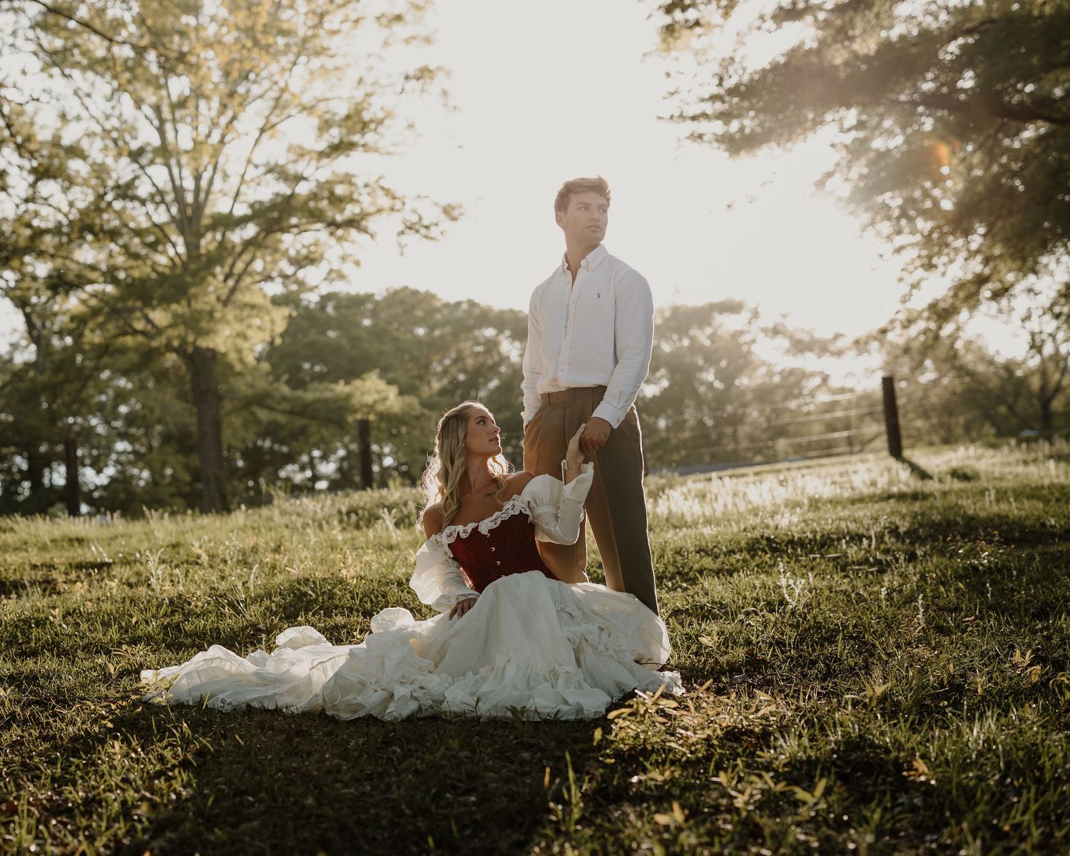 Bride in red and white gown sits on grass while groom stands beside her in golden sunset light.