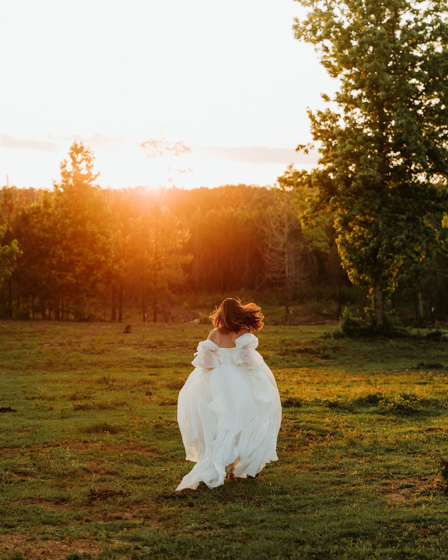 A bride in a white gown twirls in a sunlit meadow at golden hour with trees in the background.