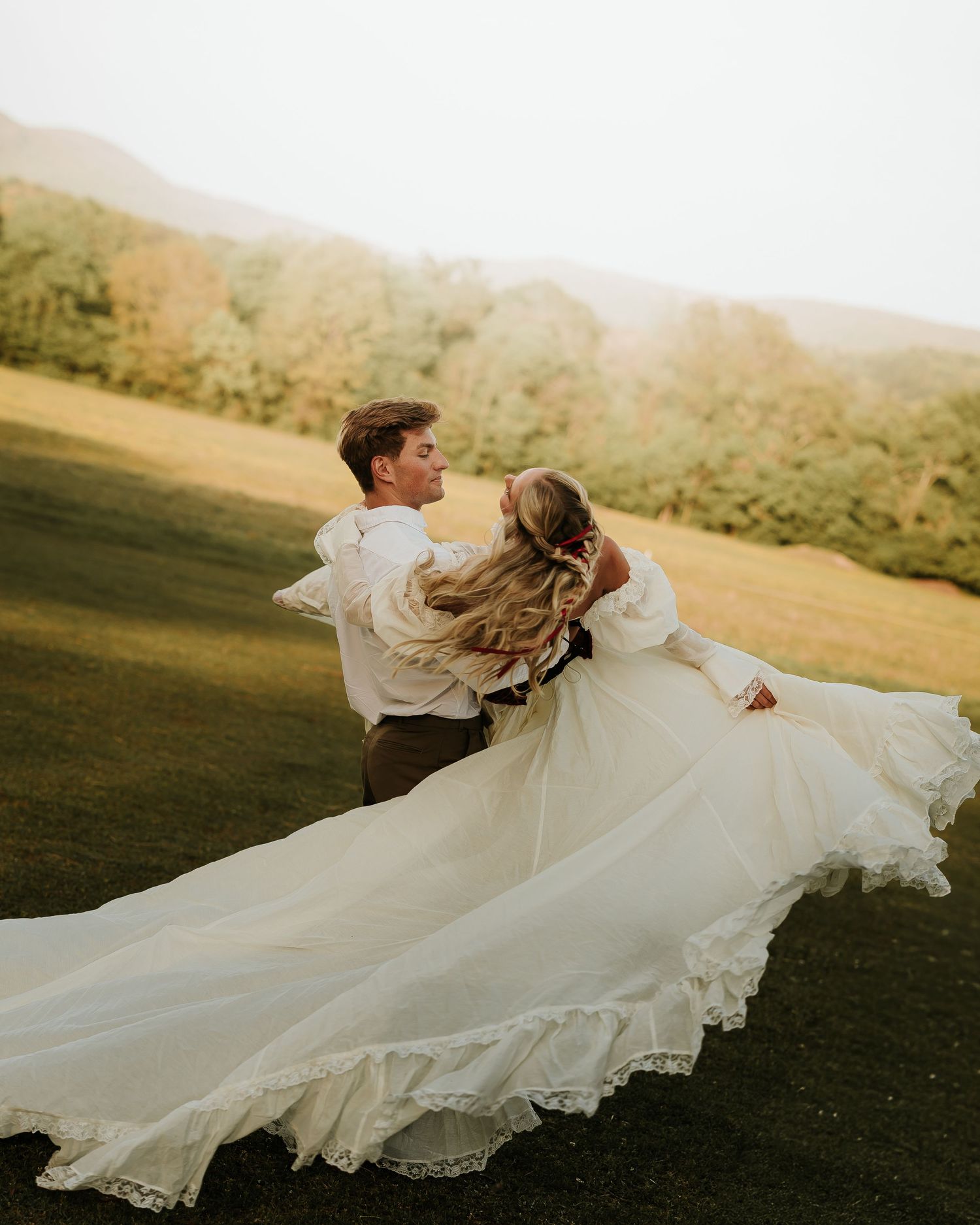 A groom twirls his bride in a flowing white wedding gown across a lush green meadow with misty mountains behind them.