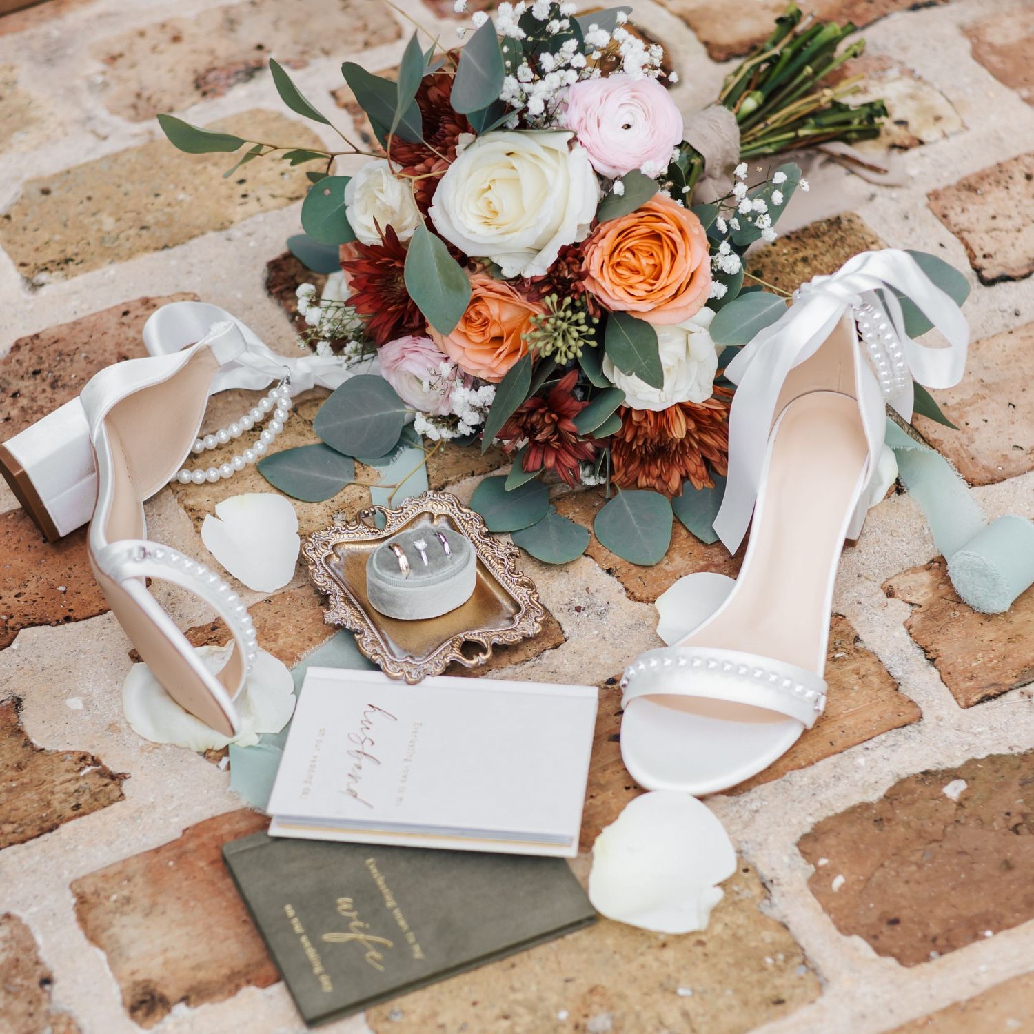 Wedding accessories including white heels, flowers, and invitation details arranged on rustic brick surface.