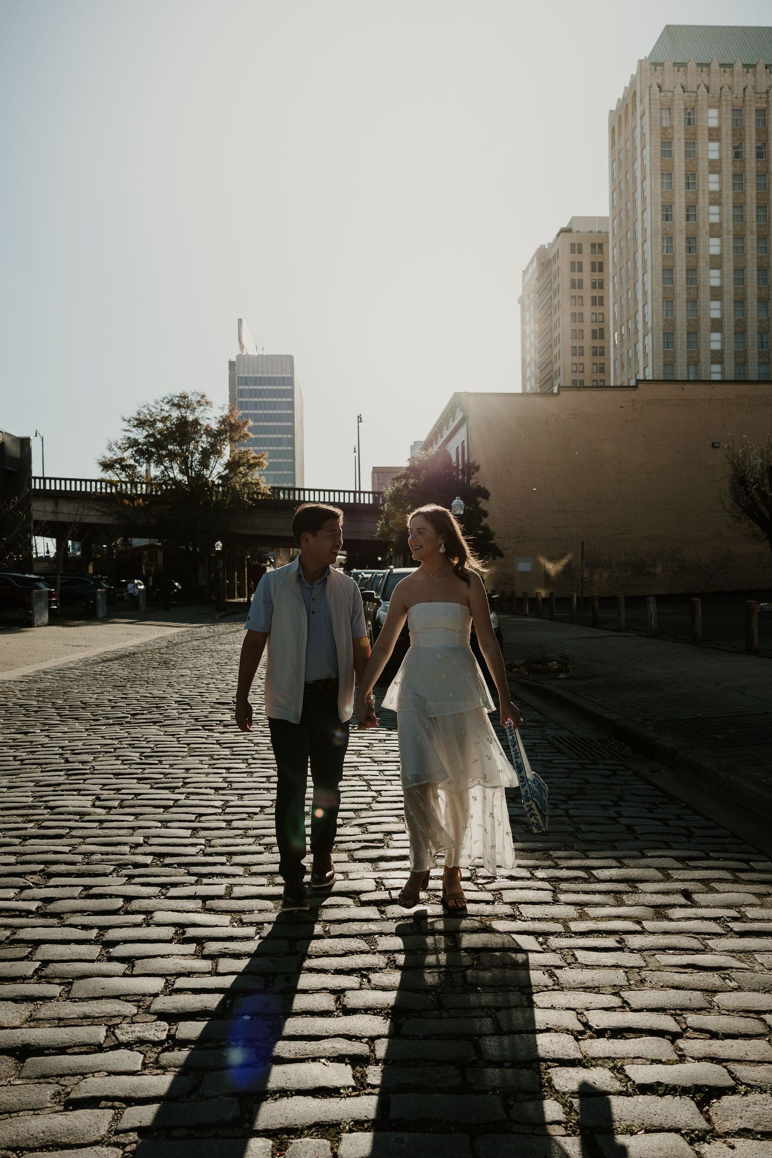 A couple walks hand in hand on cobblestone path at sunset casting long shadows between urban buildings.