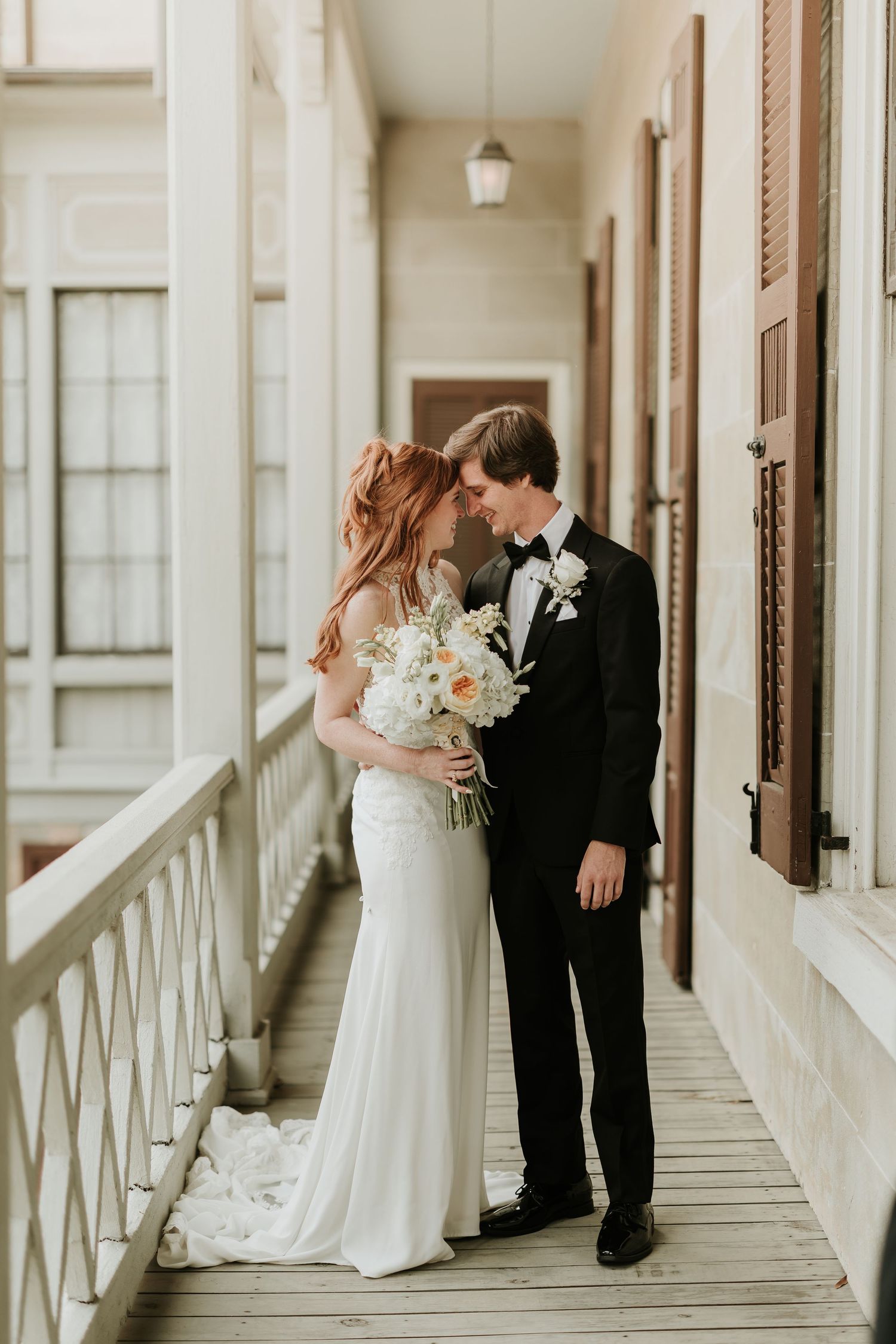 Bride and groom share an intimate moment on a historic building balcony, she holding a white bouquet in a fitted gown.
