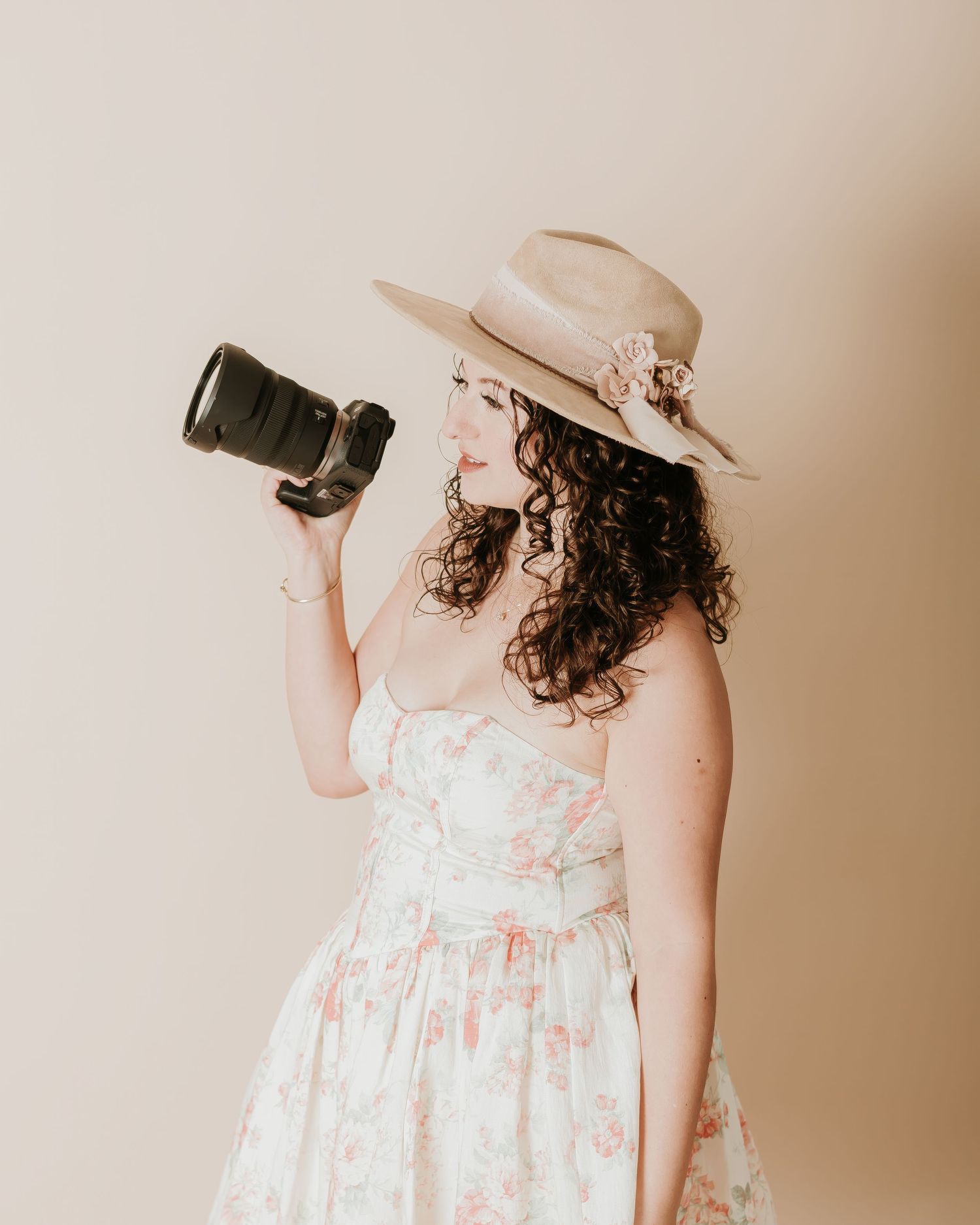 Woman in floral dress and tan hat holds DSLR camera, posed against neutral beige background.