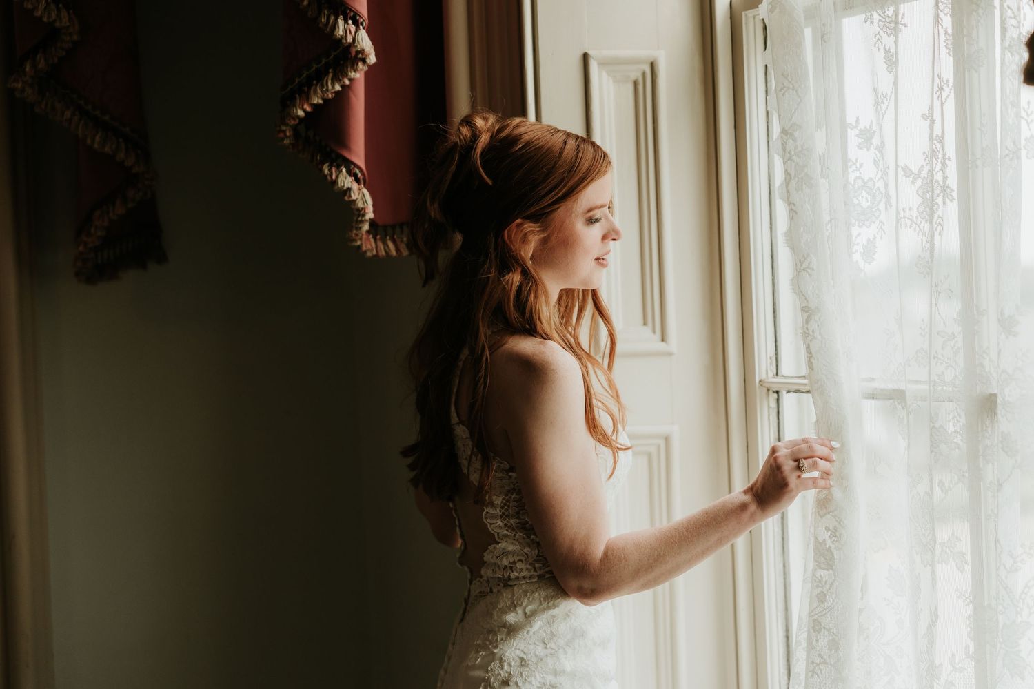A bride in a lace wedding dress gazes out a window, her auburn hair styled elegantly for her wedding day.