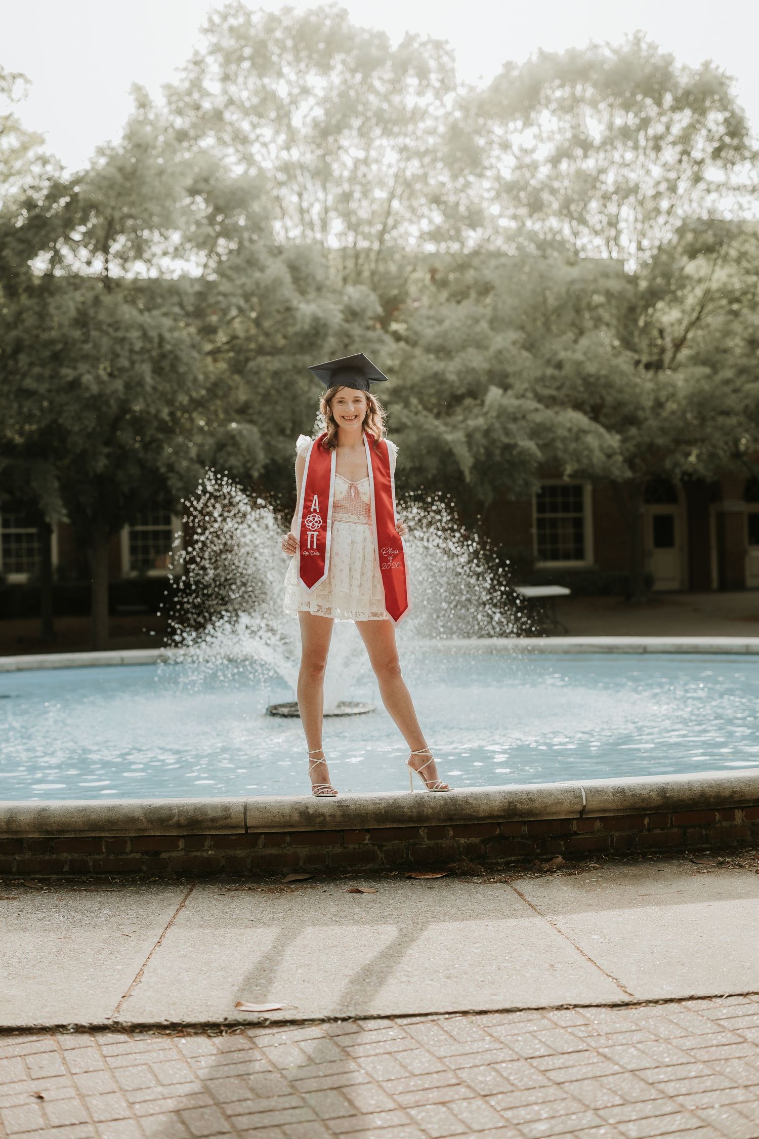 A graduate in cap and red sash poses in a fountain wearing a white dress and heels on her graduation day.