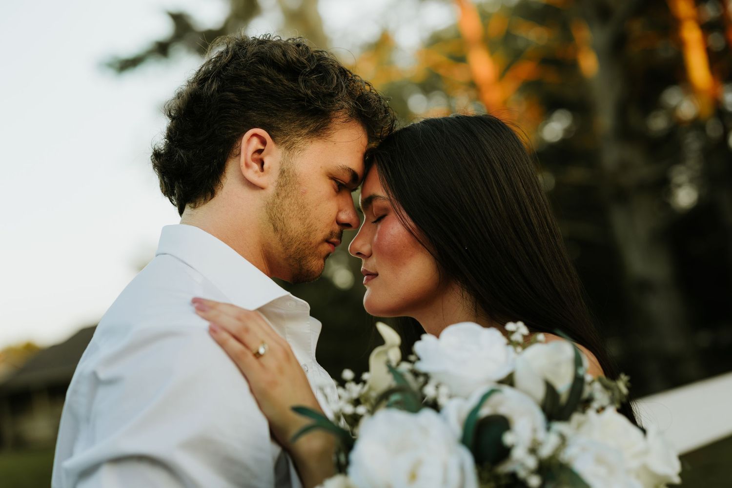 Romantic couple sharing an intimate moment at their wedding, bride holding white roses bouquet outdoors.