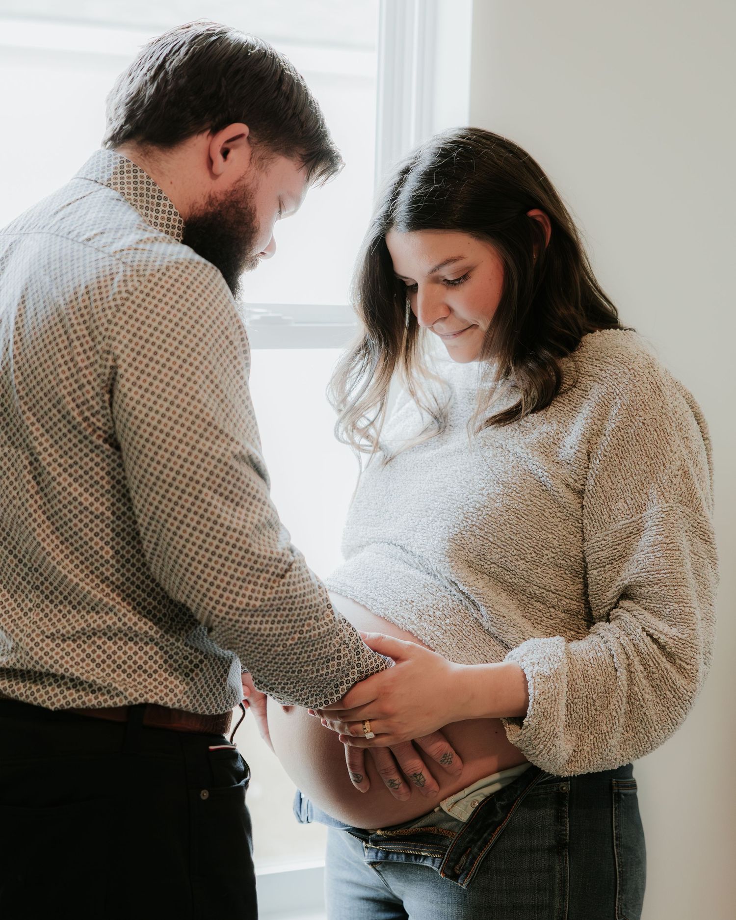 Couple shares intimate moment while touching pregnant belly by window.
