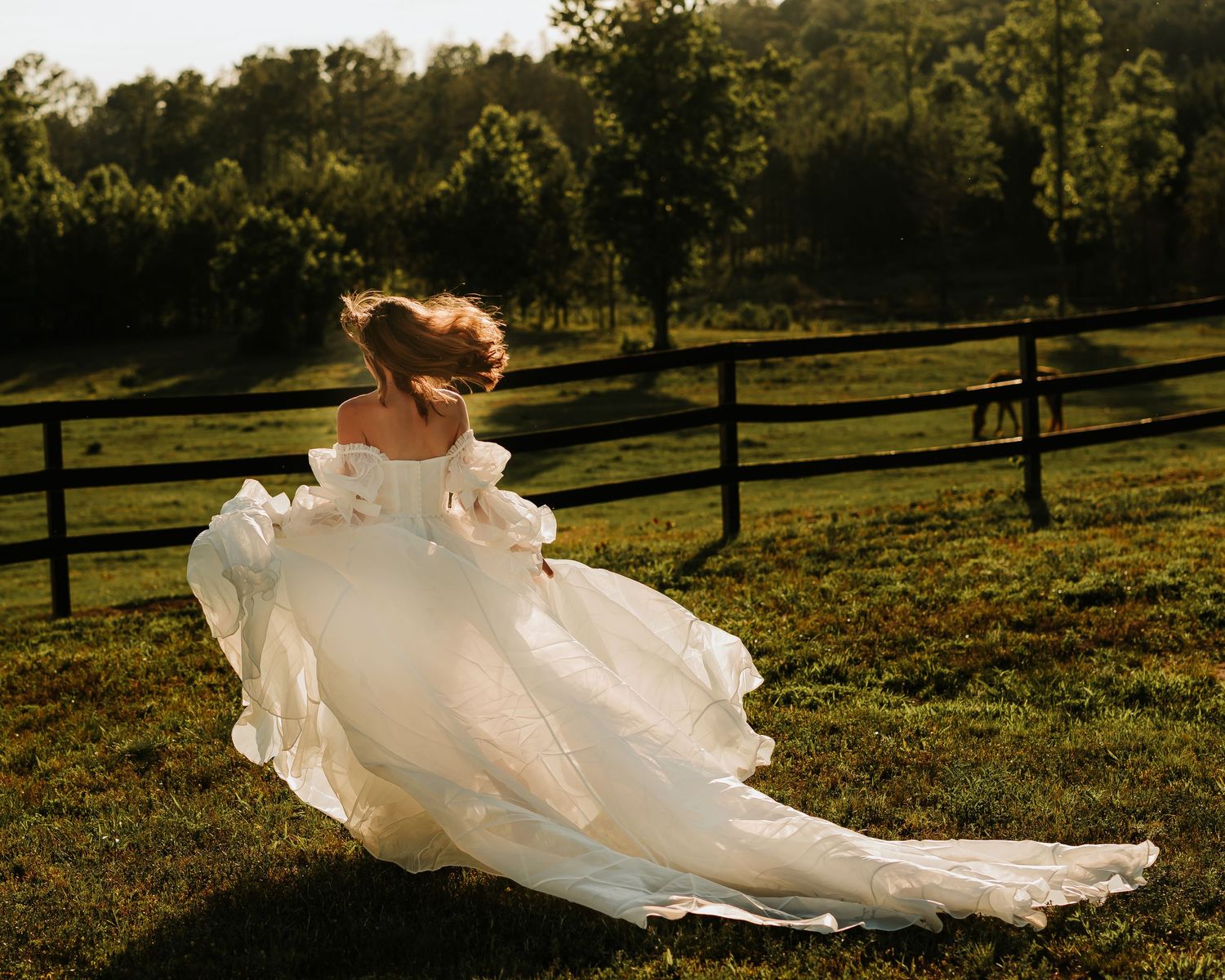 Bride in flowing white wedding gown twirling in sunlit green pasture with wooden fence and trees in background.