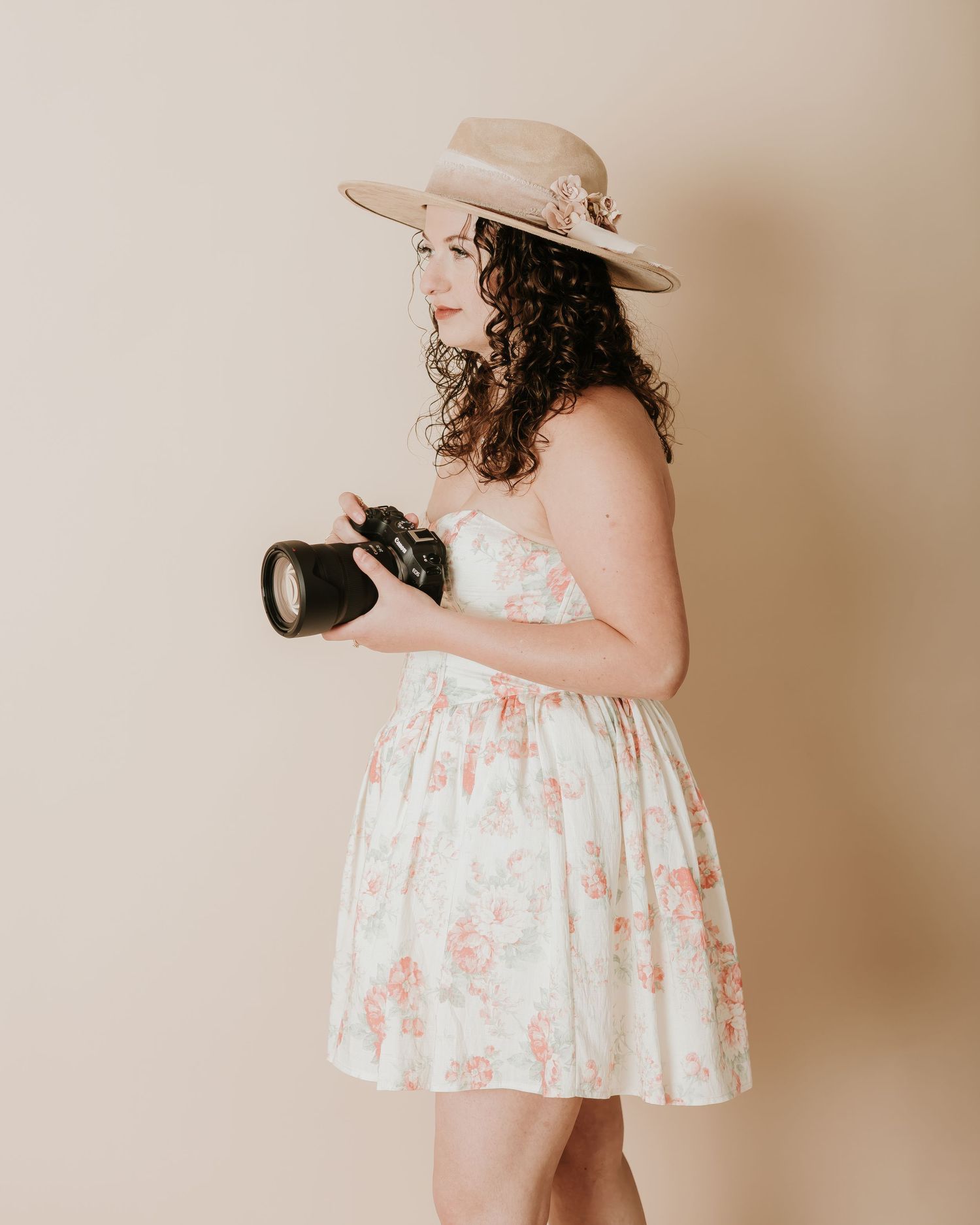 Woman in floral dress and wide-brim hat holding a DSLR camera, posing sideways against a neutral beige background.