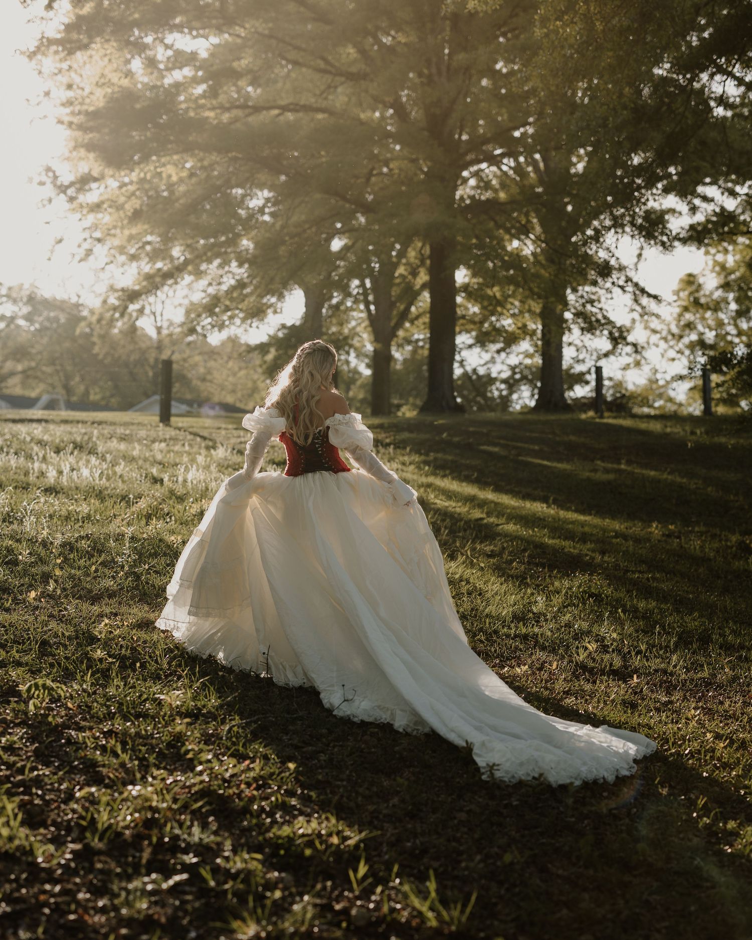 Bride twirls in flowing white wedding gown with red corset in golden hour sunlight among tall trees.