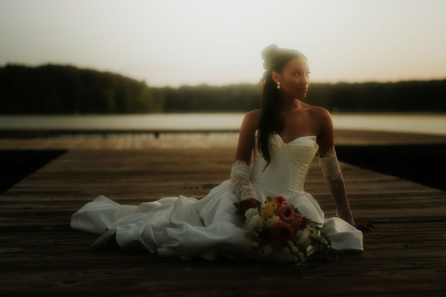 Bride in white strapless gown sitting on wooden dock at sunset holding colorful bouquet with serene lake backdrop.
