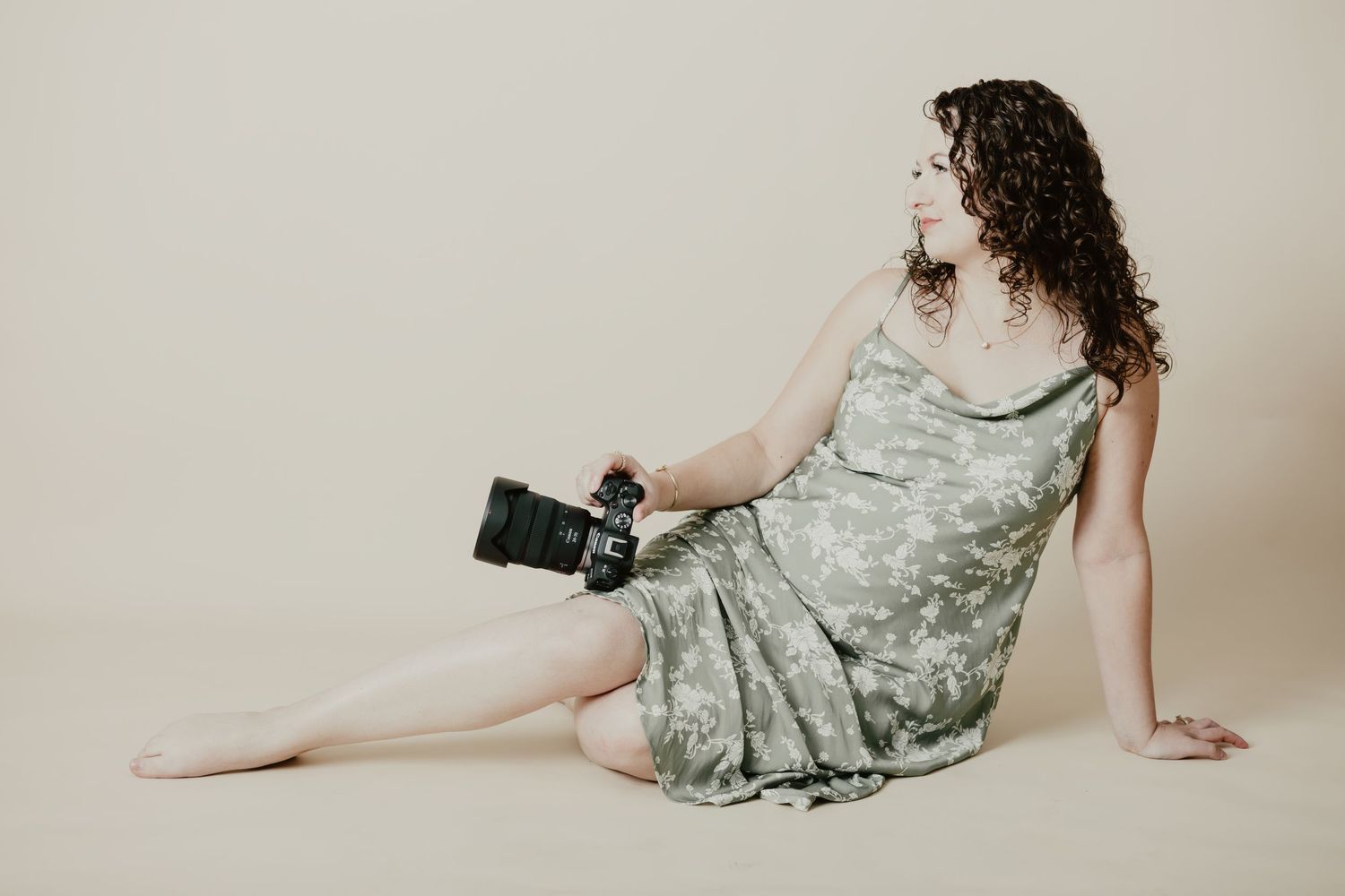 Curly-haired woman in camo dress sits on floor holding professional camera against neutral background.