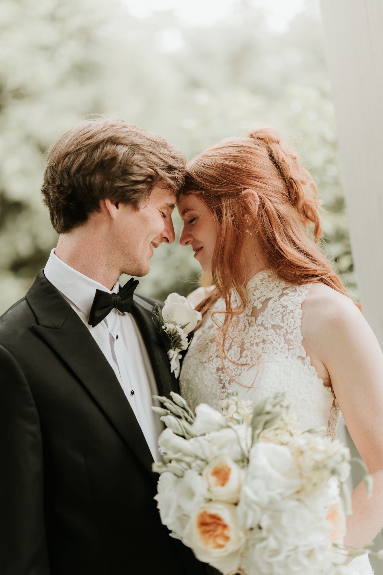 Bride and groom touching foreheads romantically, bride holding white floral bouquet, groom in black tuxedo with bow tie.
