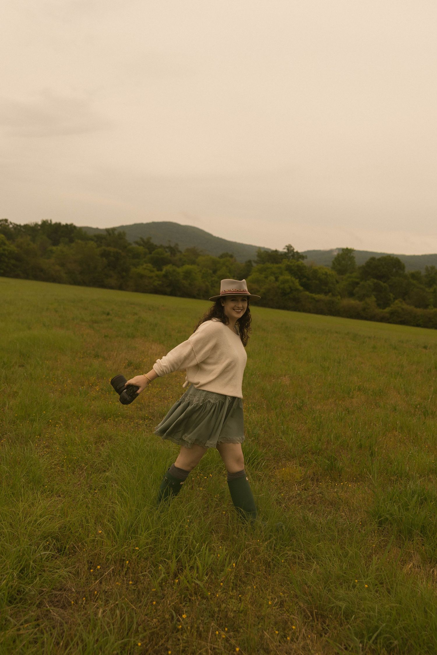 Young woman in bucket hat, white sweater, green skirt and rain boots dancing in a lush green field with mountains behind.
