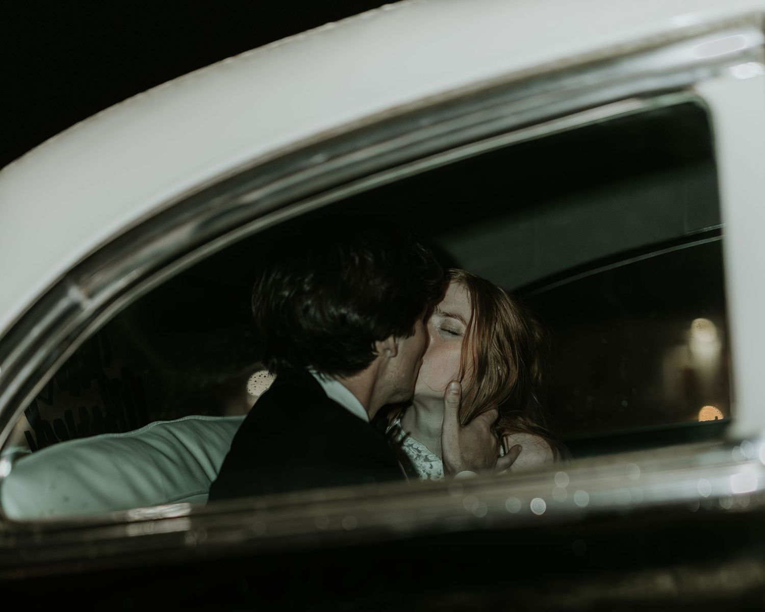 A couple shares a romantic kiss inside a vintage car at night, captured through the window.