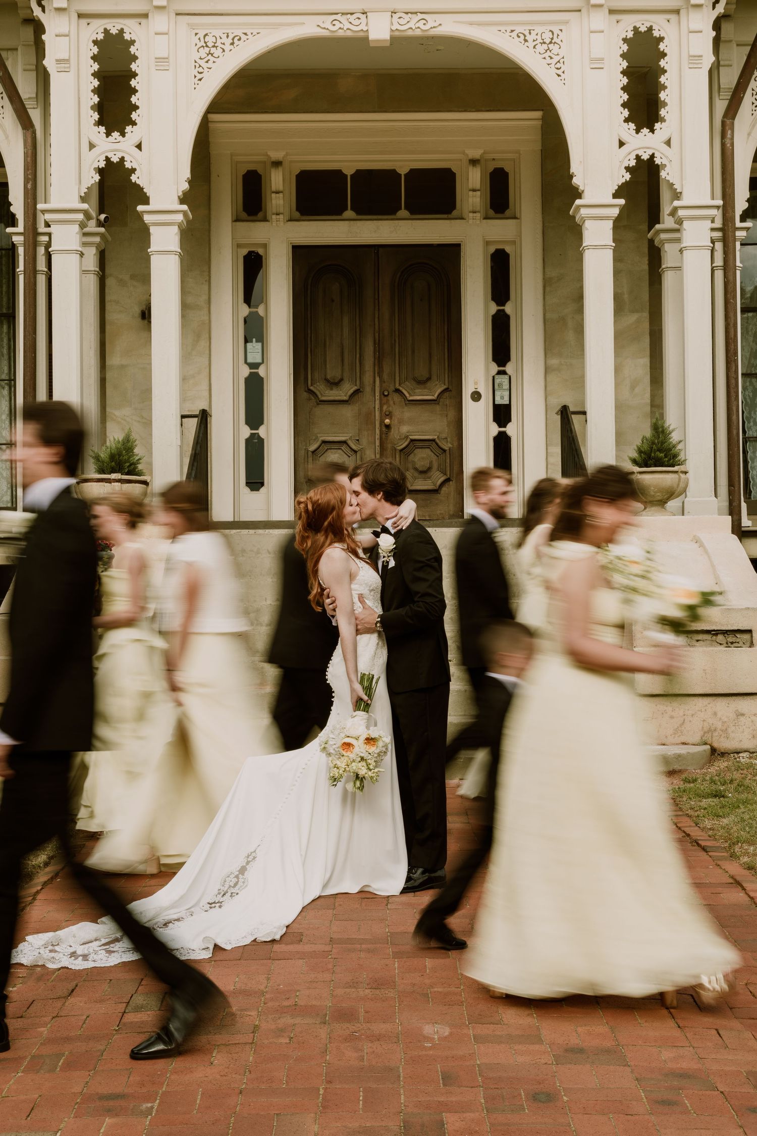 Bride and groom share a kiss while wedding party walks past in motion blur outside elegant historic venue.