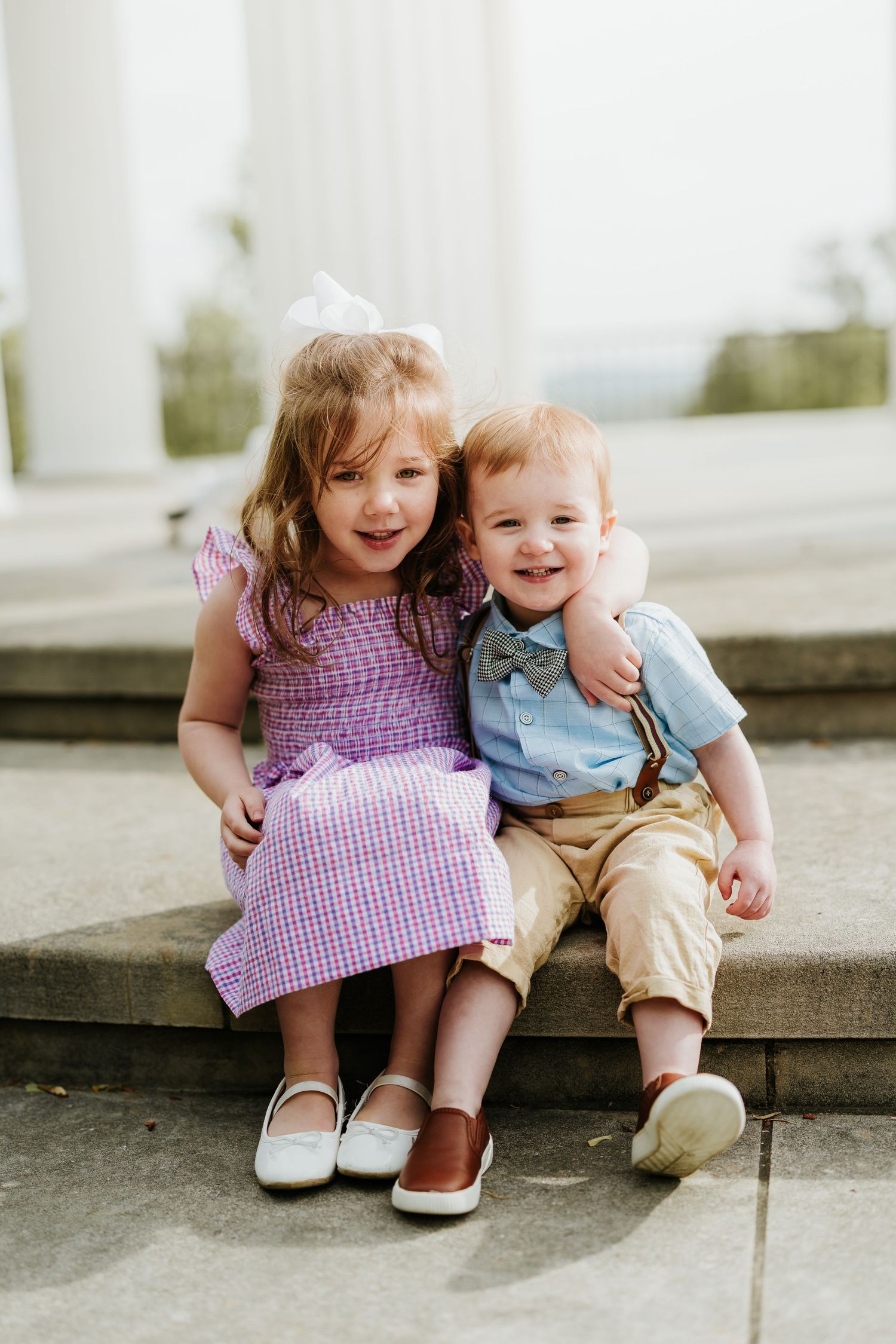 Two young siblings, a girl in purple dress and boy in blue shirt, sitting together on steps smiling.