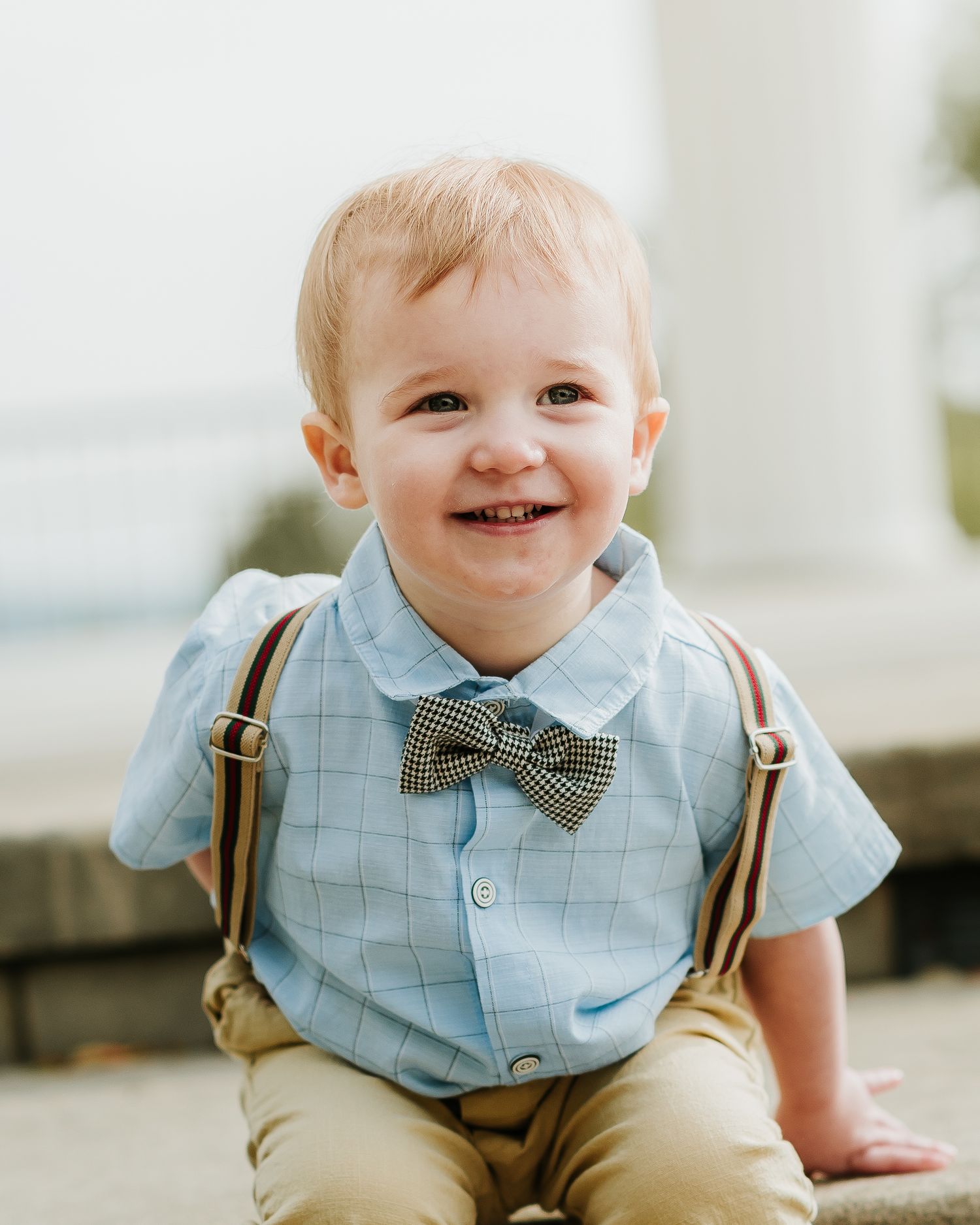 Smiling toddler boy wearing a blue plaid shirt, bow tie, and suspenders sitting outdoors for a portrait photo.
