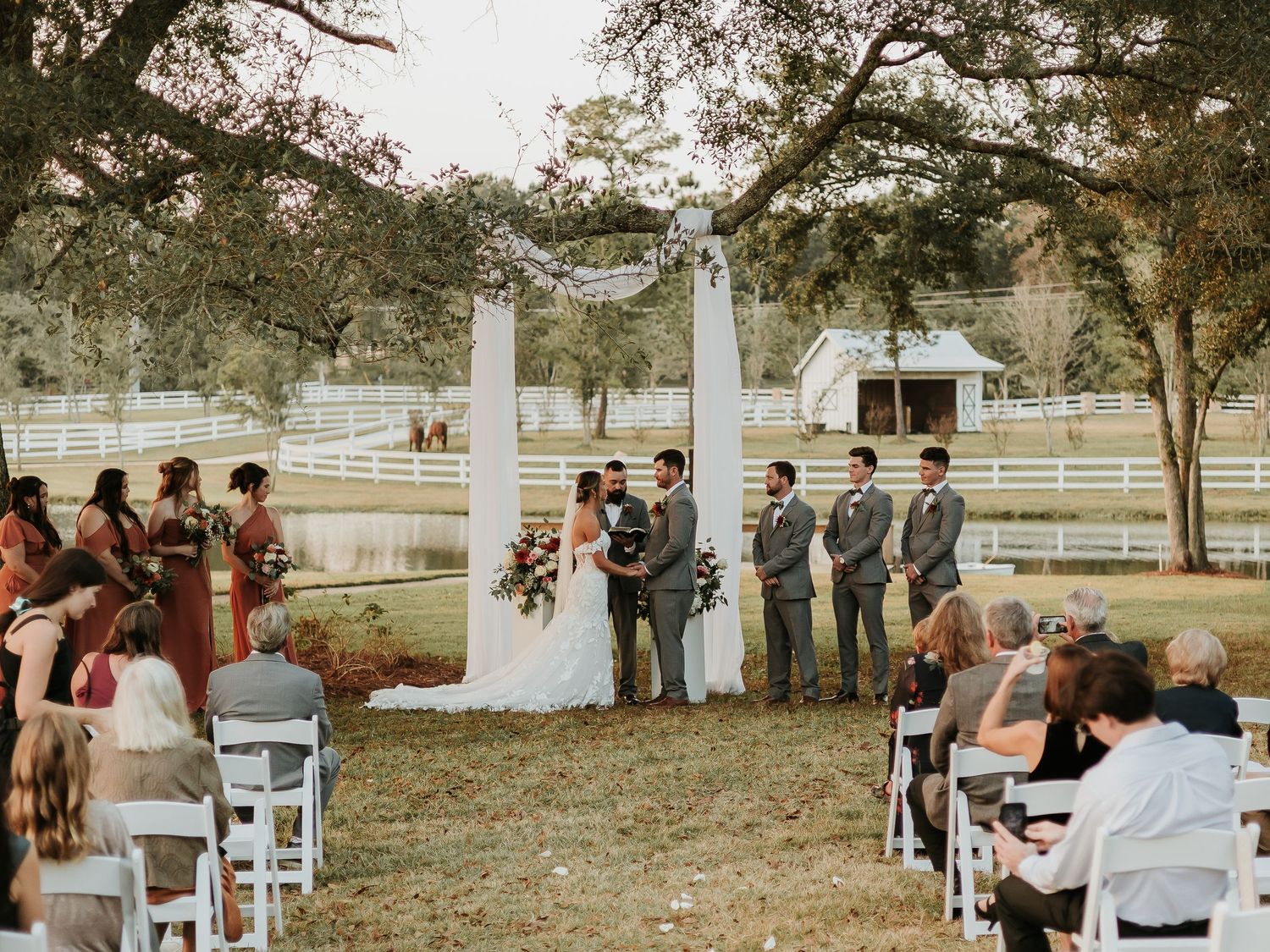 Outdoor wedding ceremony under oak trees at a rustic farm venue with white chairs, wedding party, and white fence backdrop.