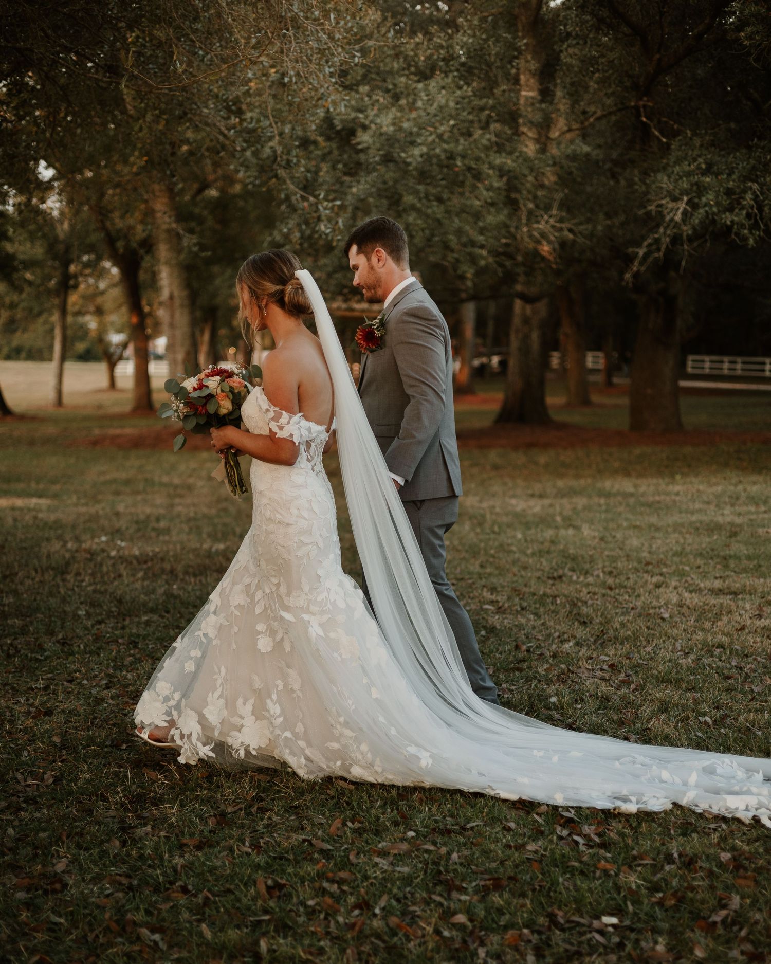 Bride in lace mermaid gown with veil and groom in gray suit share romantic moment outdoors among trees.