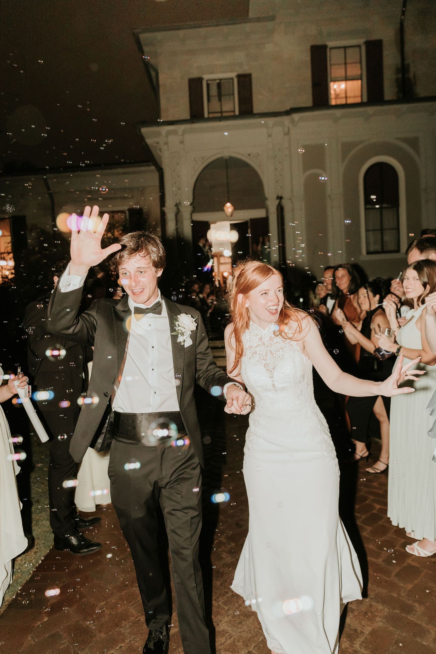 Joyful newlyweds exit wedding venue through bubble send-off, bride in white gown and groom in black tuxedo.