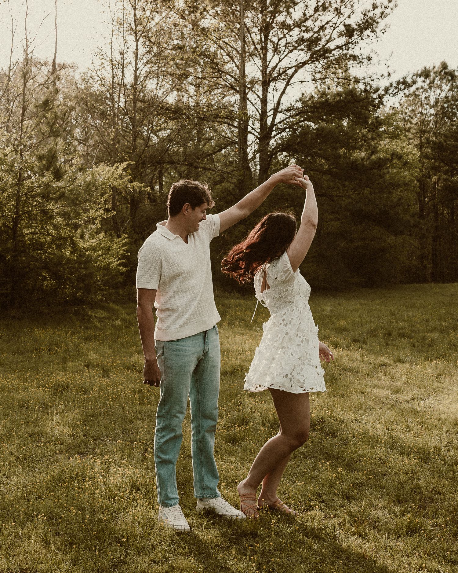 A couple dances together in a sunny green meadow, the man spinning the woman in a white floral dress.