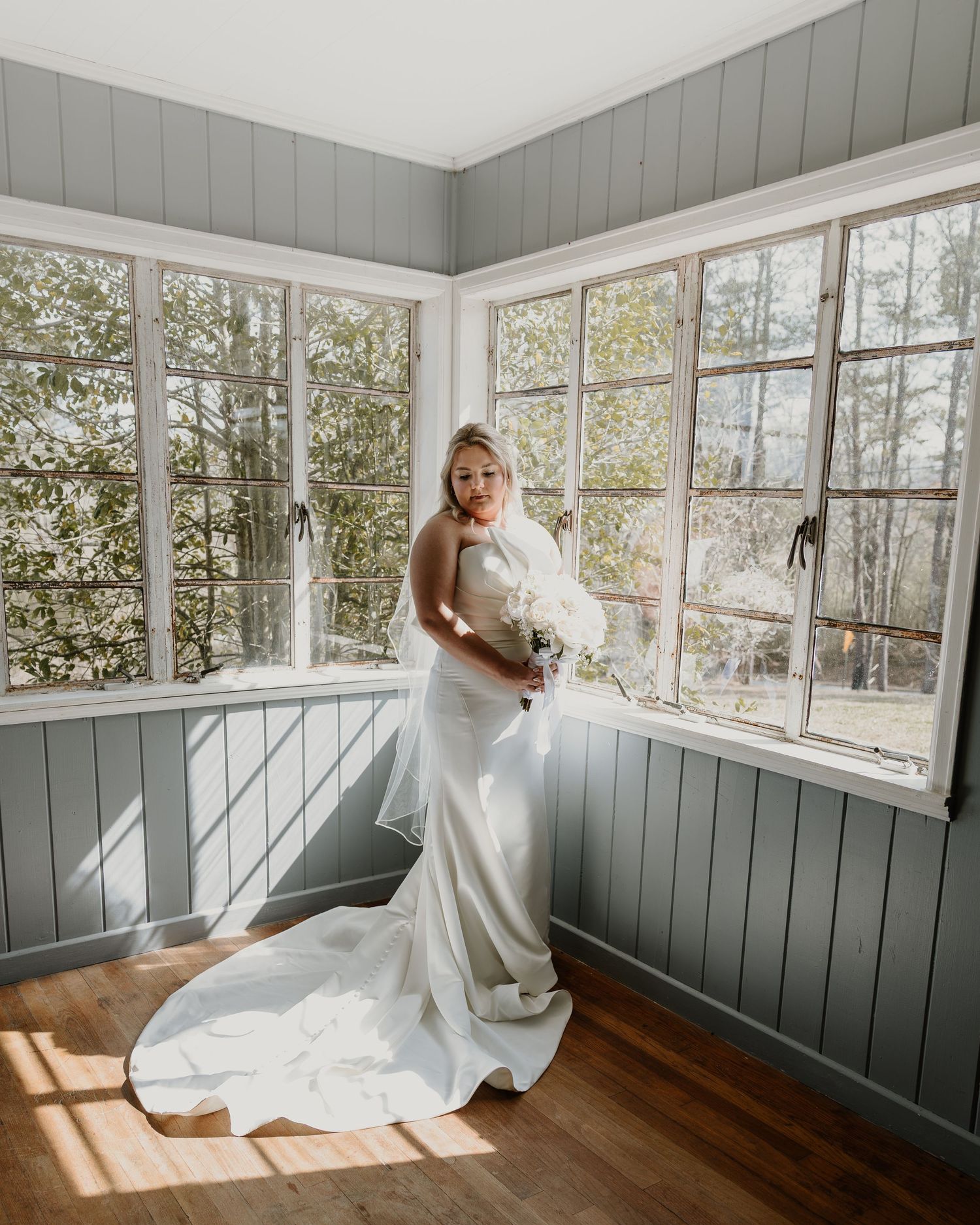 Bride in white satin gown with train holds bouquet in sunlit room with large windows and wood floors.