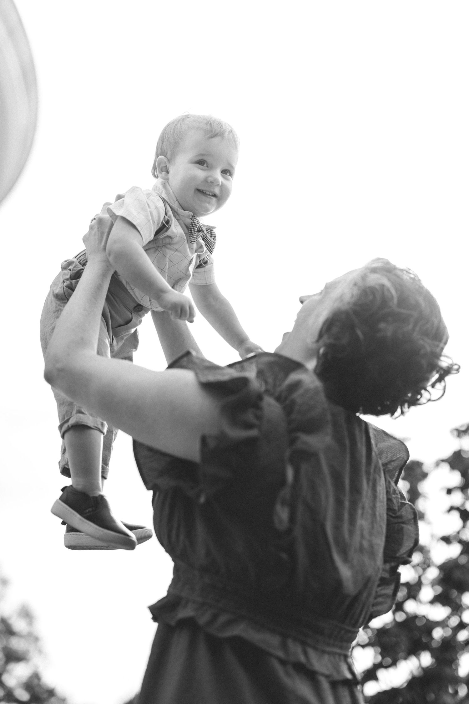 Black and white photo of mother lifting smiling baby into the air outdoors against bright sky.