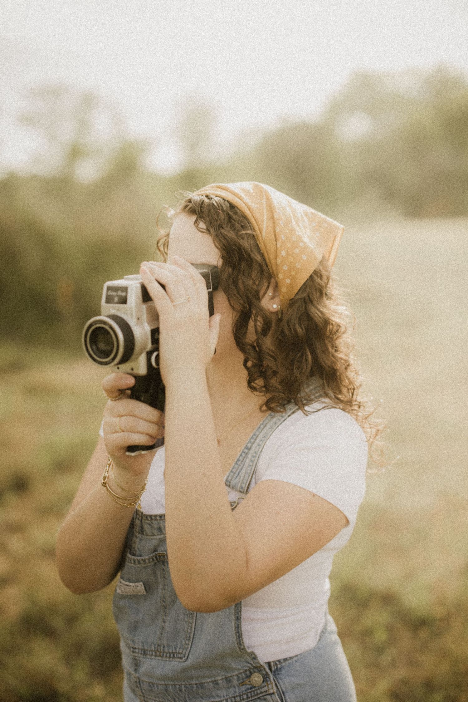 Young woman in denim overalls and bandana holding a vintage film camera outdoors in a golden field.