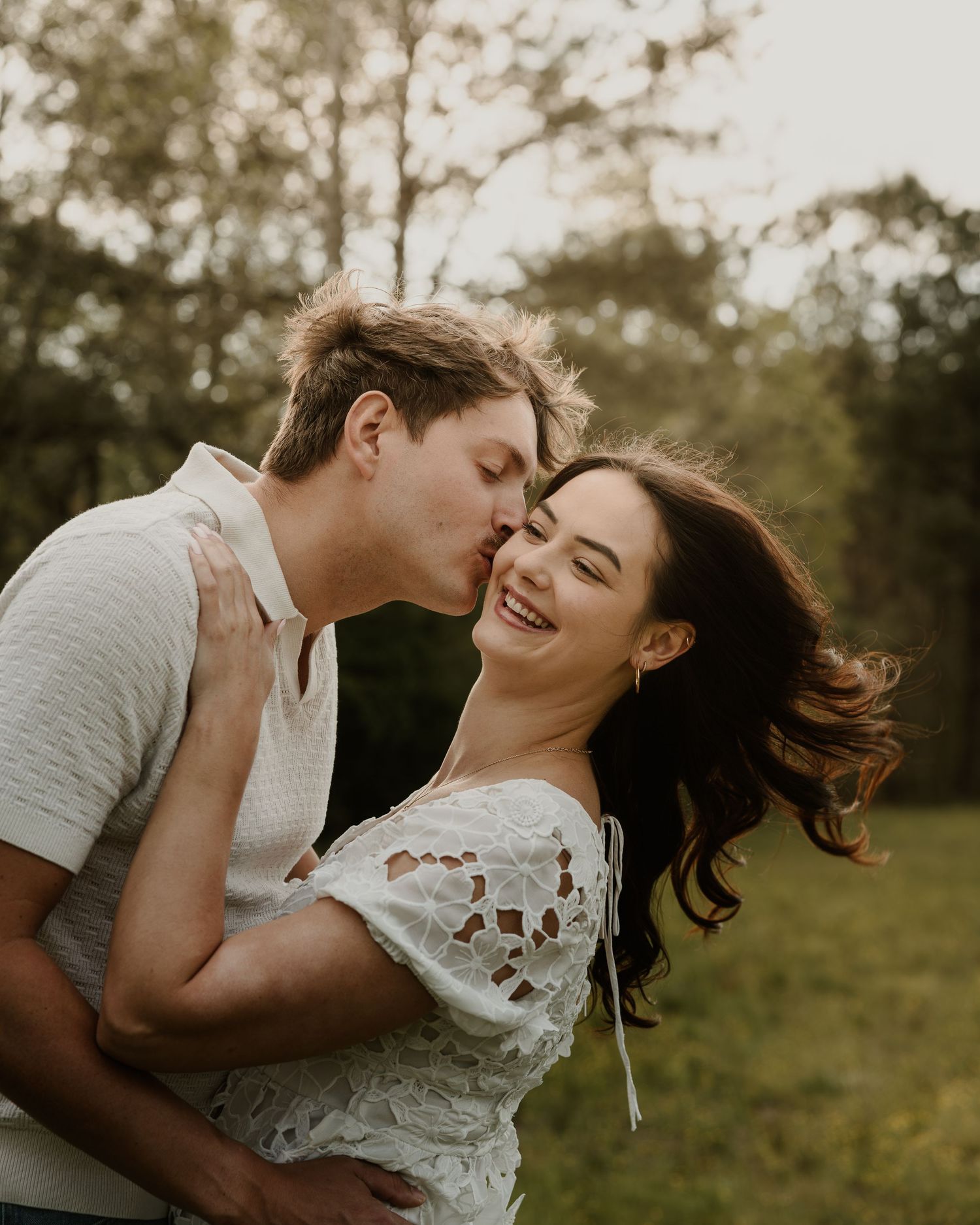 A couple shares a romantic kiss outdoors, with the woman smiling joyfully in a white lace top amid lush greenery.