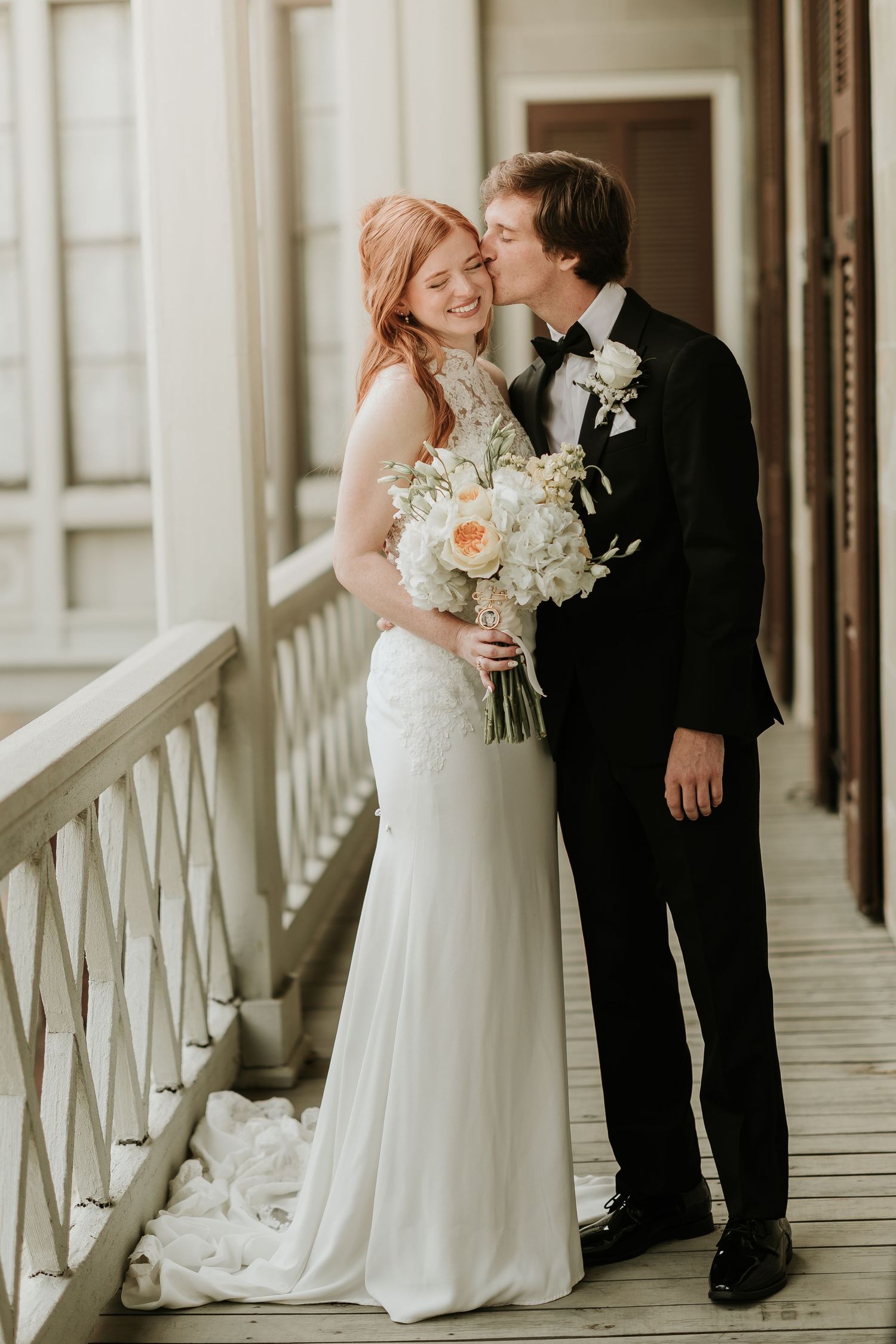 Bride in white gown holds floral bouquet as groom in black tuxedo kisses her cheek on a elegant balcony.