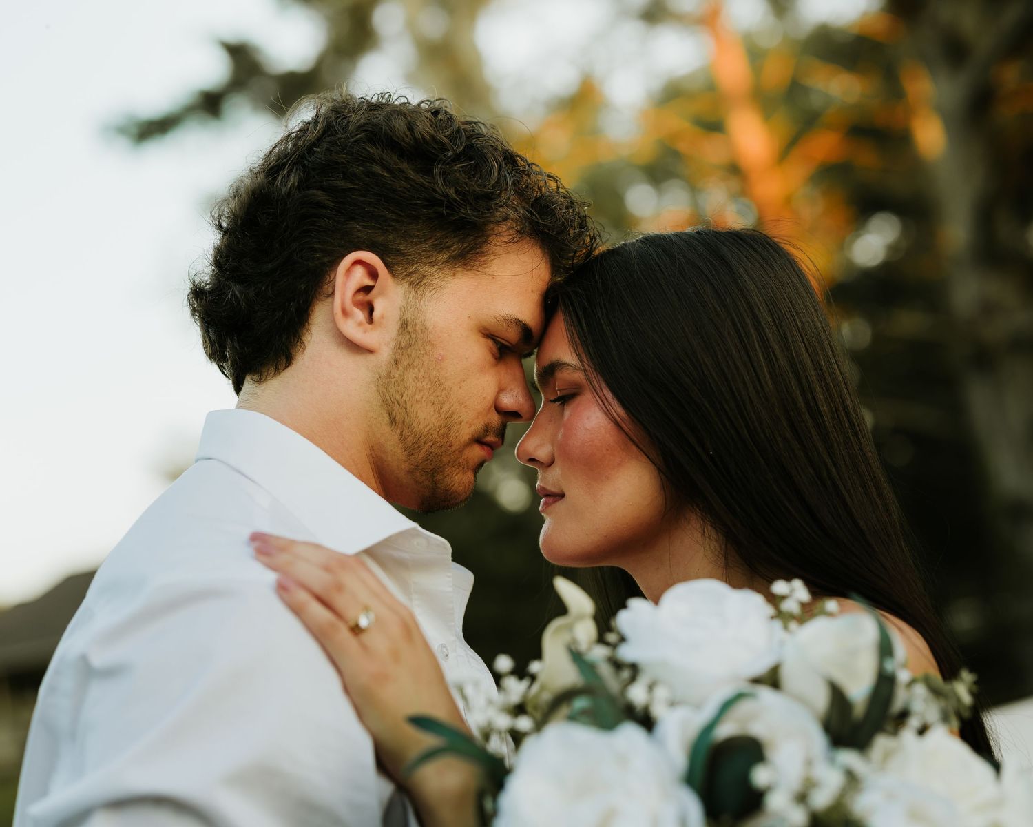 A romantic couple in wedding attire share an intimate forehead-to-forehead moment, bride holding white floral bouquet outdoors.