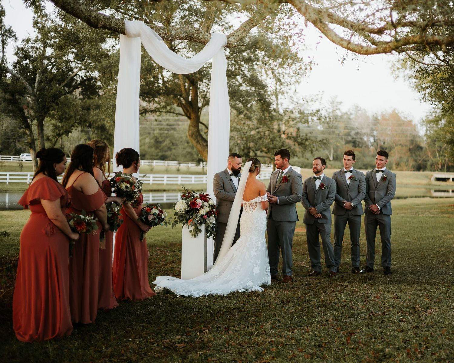 Wedding party stands under white arch outdoors with bridesmaids in rust dresses and groomsmen in gray suits.