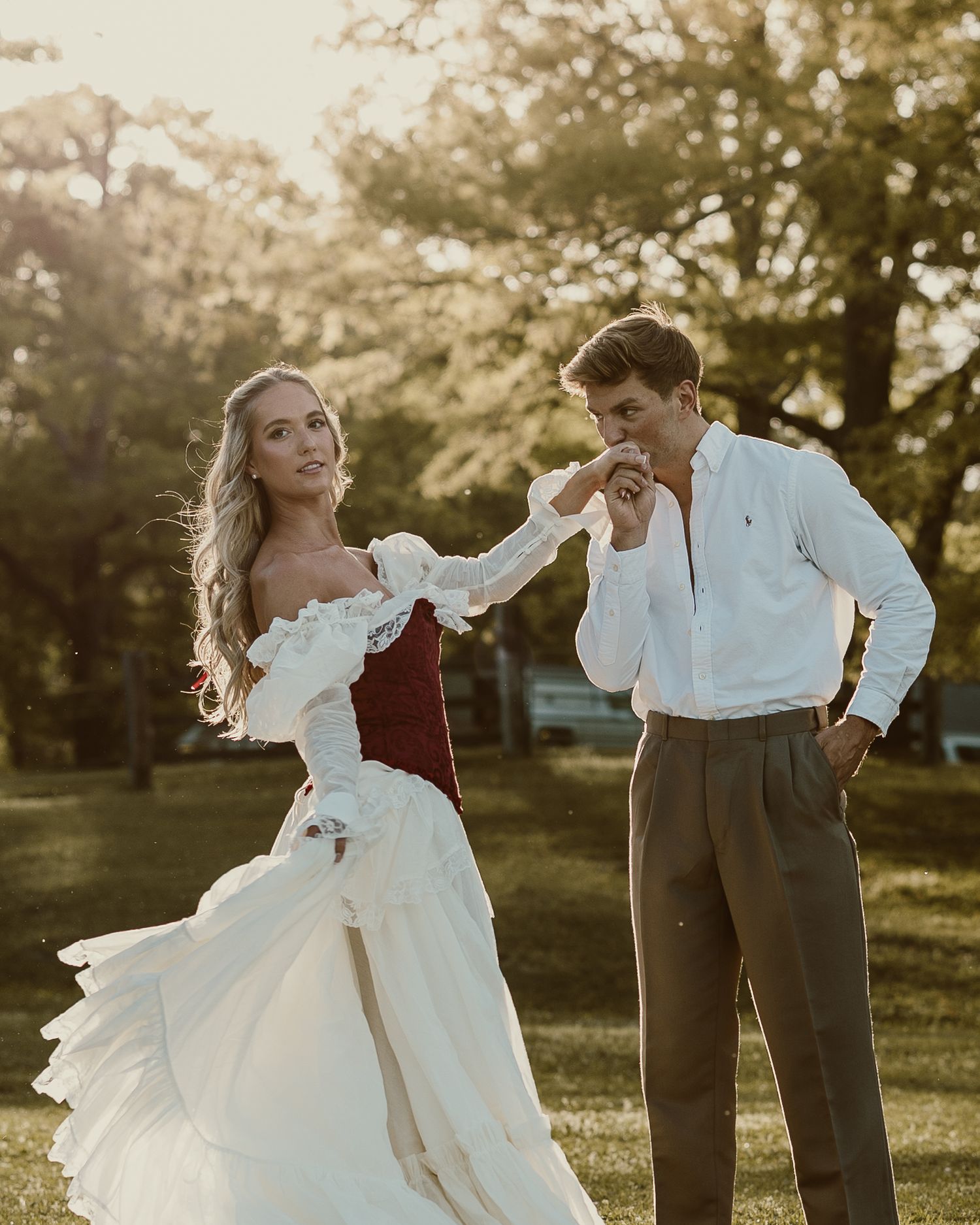 A couple in elegant attire dances outdoors at golden hour, man kissing womans hand in white and burgundy gown.