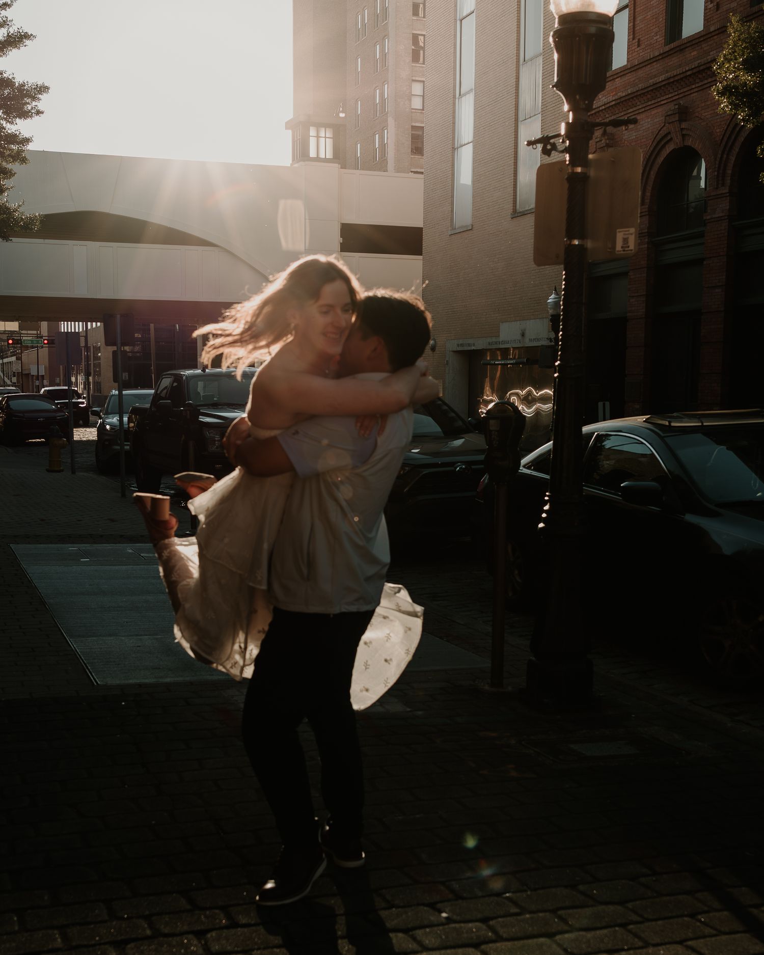 A romantic silhouette of a couple embracing on a city street at sunset with sunlight streaming between buildings.