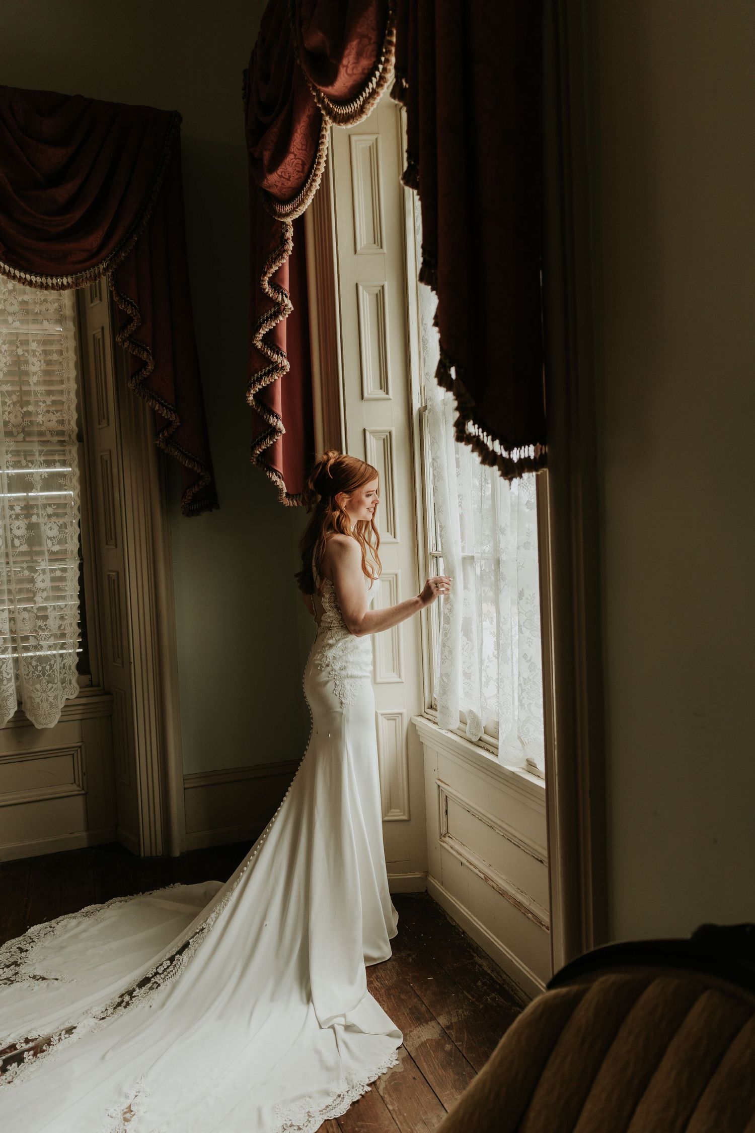 Bride in white mermaid gown with lace train stands by tall window with red drapes in elegant vintage room.