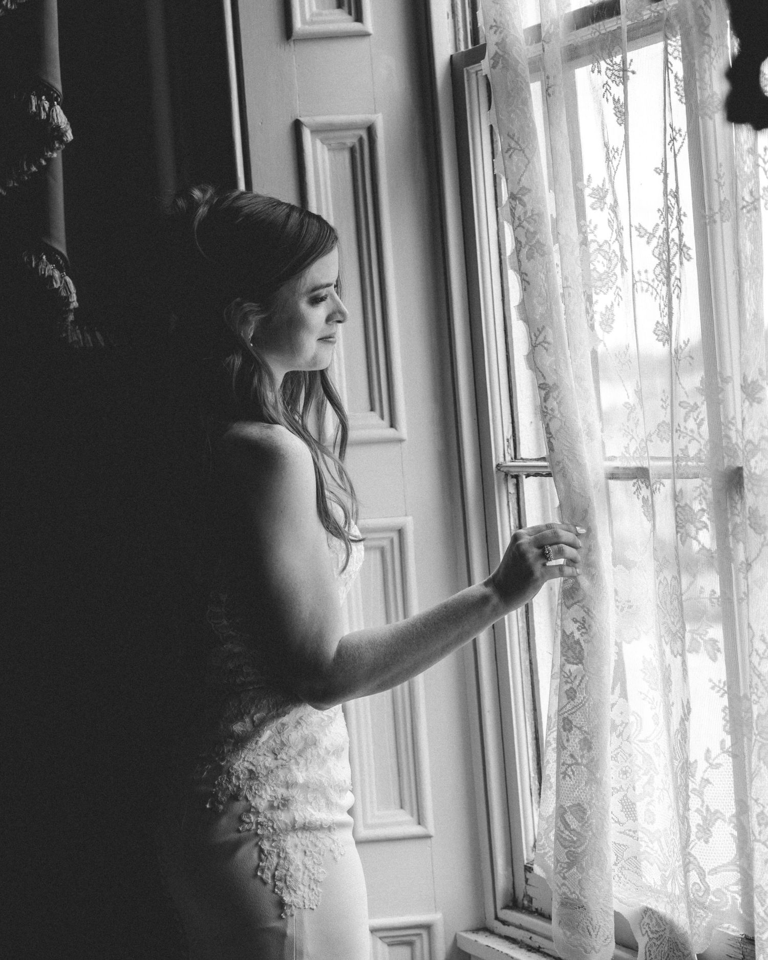 Black and white photo of a bride in a lace gown gazing out a window, holding sheer curtains.