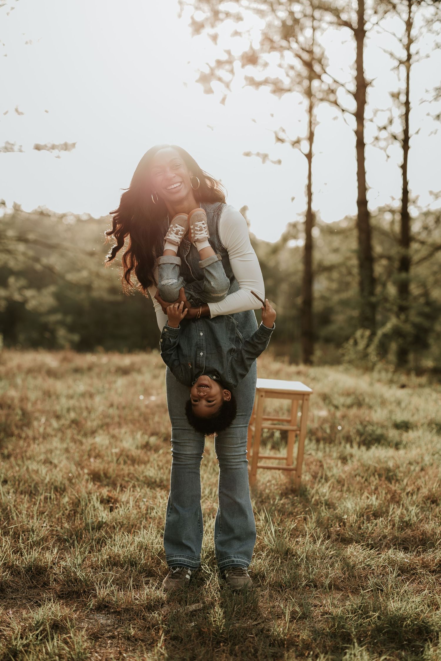 Man playfully holding woman upside down while laughing together in a sunlit forest meadow.