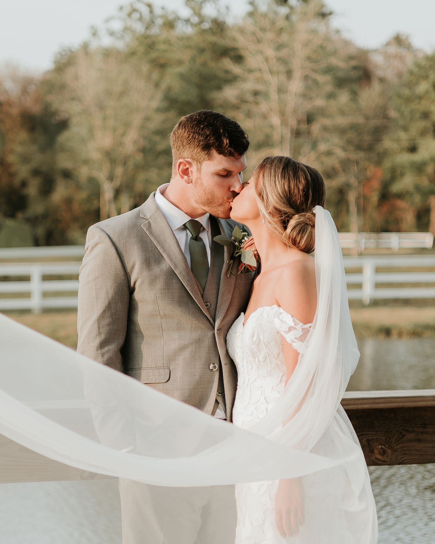 Bride and groom share a romantic kiss outdoors, with her veil flowing in the breeze at golden hour.