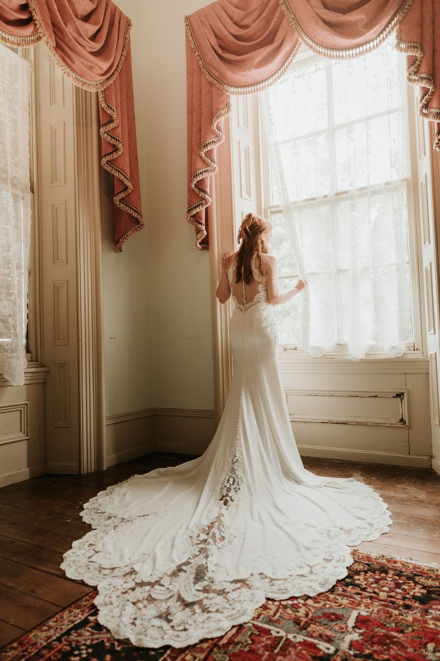 Bride in a elegant lace-train wedding gown stands by a sunlit window with pink drapes in a classic interior.