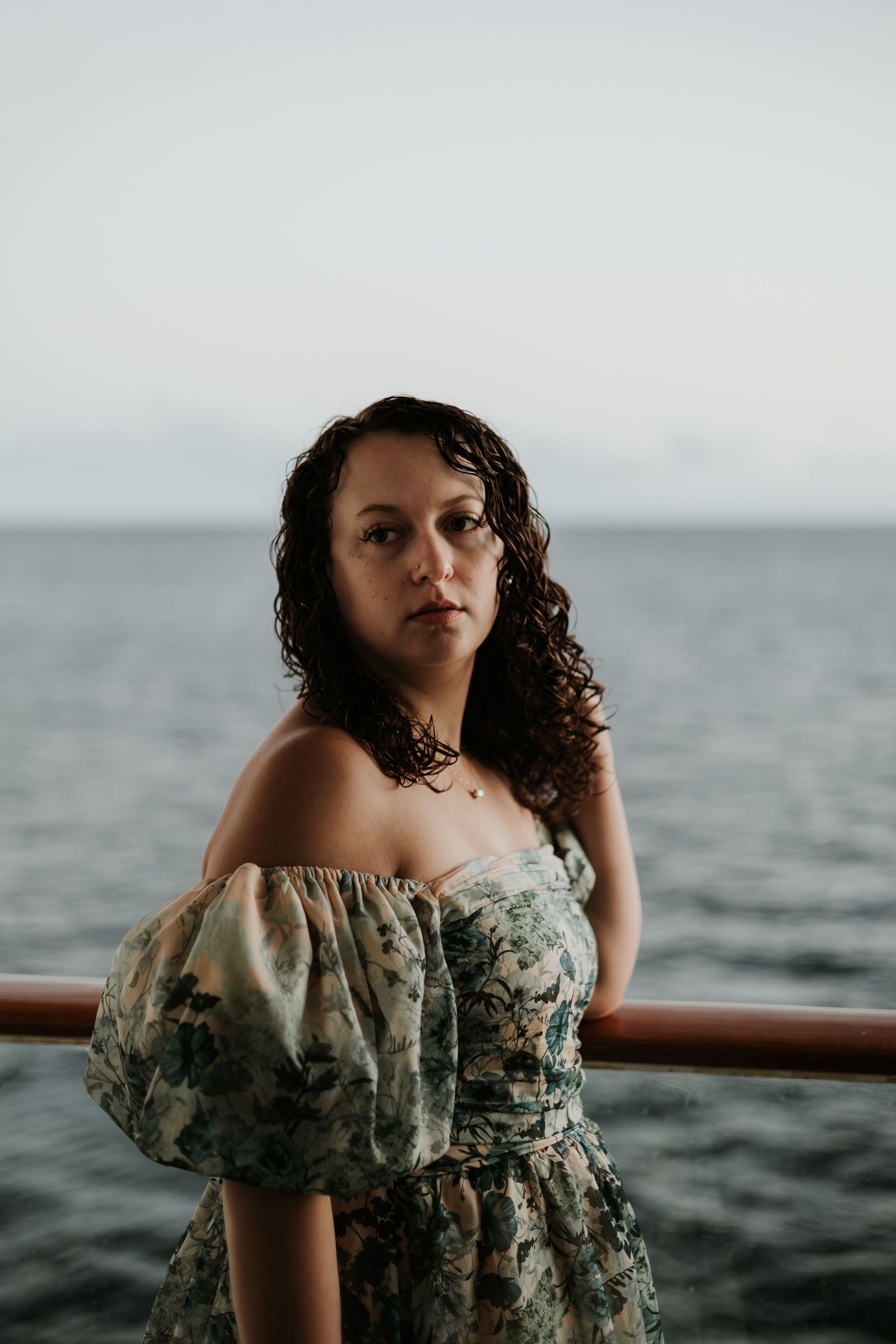 A young woman with curly hair in a floral off-shoulder dress poses by a railing with ocean views behind her.