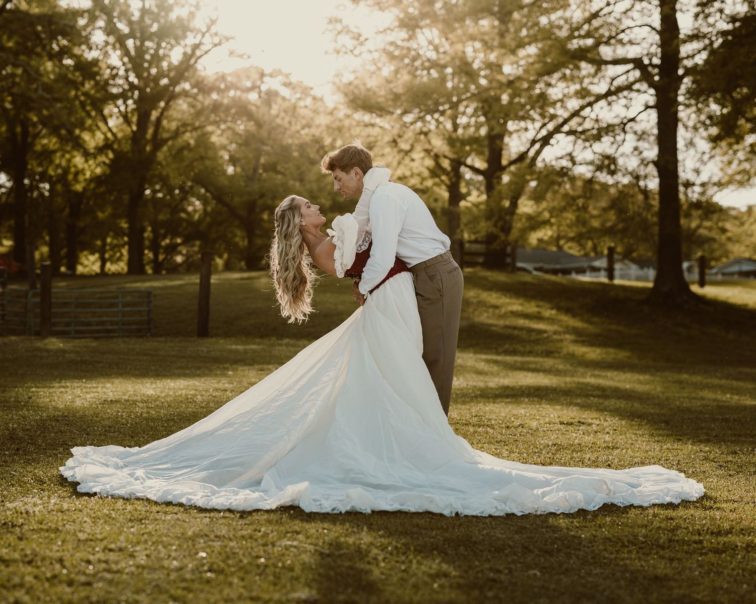 Bride and groom share a romantic dip kiss on lush green grass at golden hour, with trees glowing in warm sunlight.